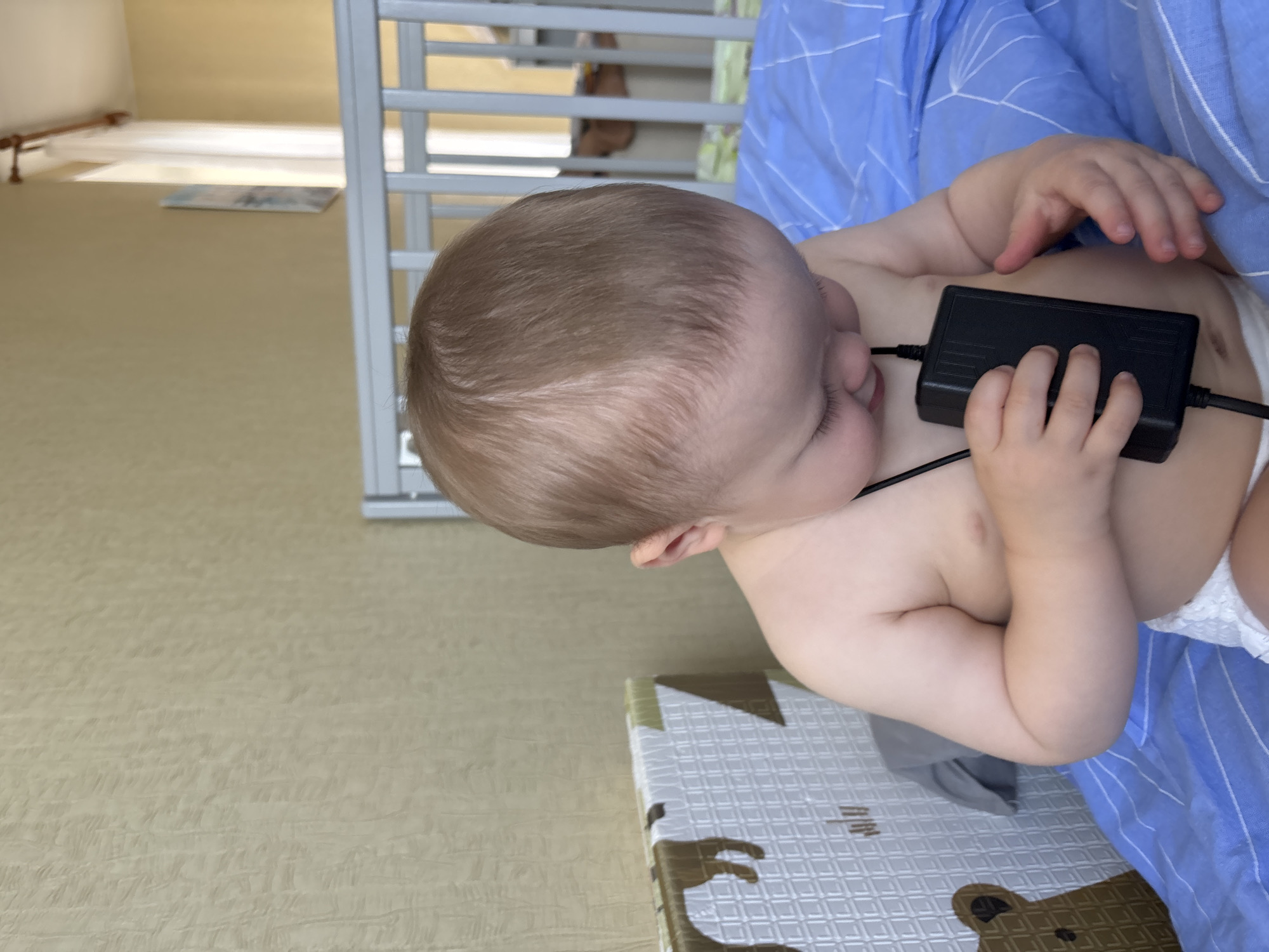 A close-up, high-angle shot shows a baby lying on its back. The baby has light brown hair and is wearing a diaper. The baby's head is turned to the left, and its eyes are closed. A black power adapter is placed on the baby's chest, with cords extending to the sides. The baby's hands are positioned around the adapter, with one hand resting on top of it. The baby is lying on a blue and white striped blanket, which covers the lower half of the image. The background is blurred, but appears to be an indoor setting with a crib and a rug. The lighting suggests it is daytime. The overall mood is peaceful and innocent, with the baby appearing to be asleep or resting.