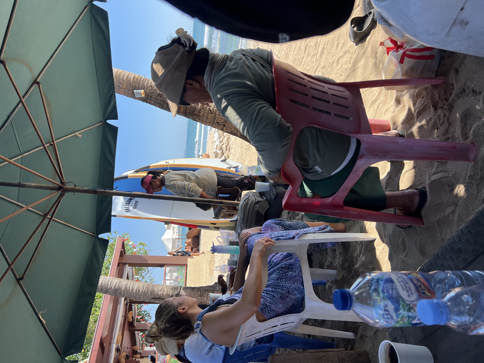 The image depicts a beach scene in Kuta, Indonesia, likely near a surf school given the presence of surfboards. In the foreground, two individuals are seated under a large green umbrella, which is angled to provide shade. The person on the left, a woman with her hair tied up, wears a patterned dress and is facing away from the camera. The person on the right, wearing a cap and sunglasses, is facing to the left, their gaze directed out towards the ocean. Both are seated in chairs, one white and one red.
Various objects are scattered around them, including water bottles, a white paper cup containing a dark liquid, and a bag with red ribbon details. In the background, more surfboards are visible, leaning against a structure. Beyond that, the ocean stretches out with small waves, and people can be seen enjoying the beach further down. Palm trees are also present, adding to the tropical atmosphere. The overall scene suggests a relaxed, beachside environment.