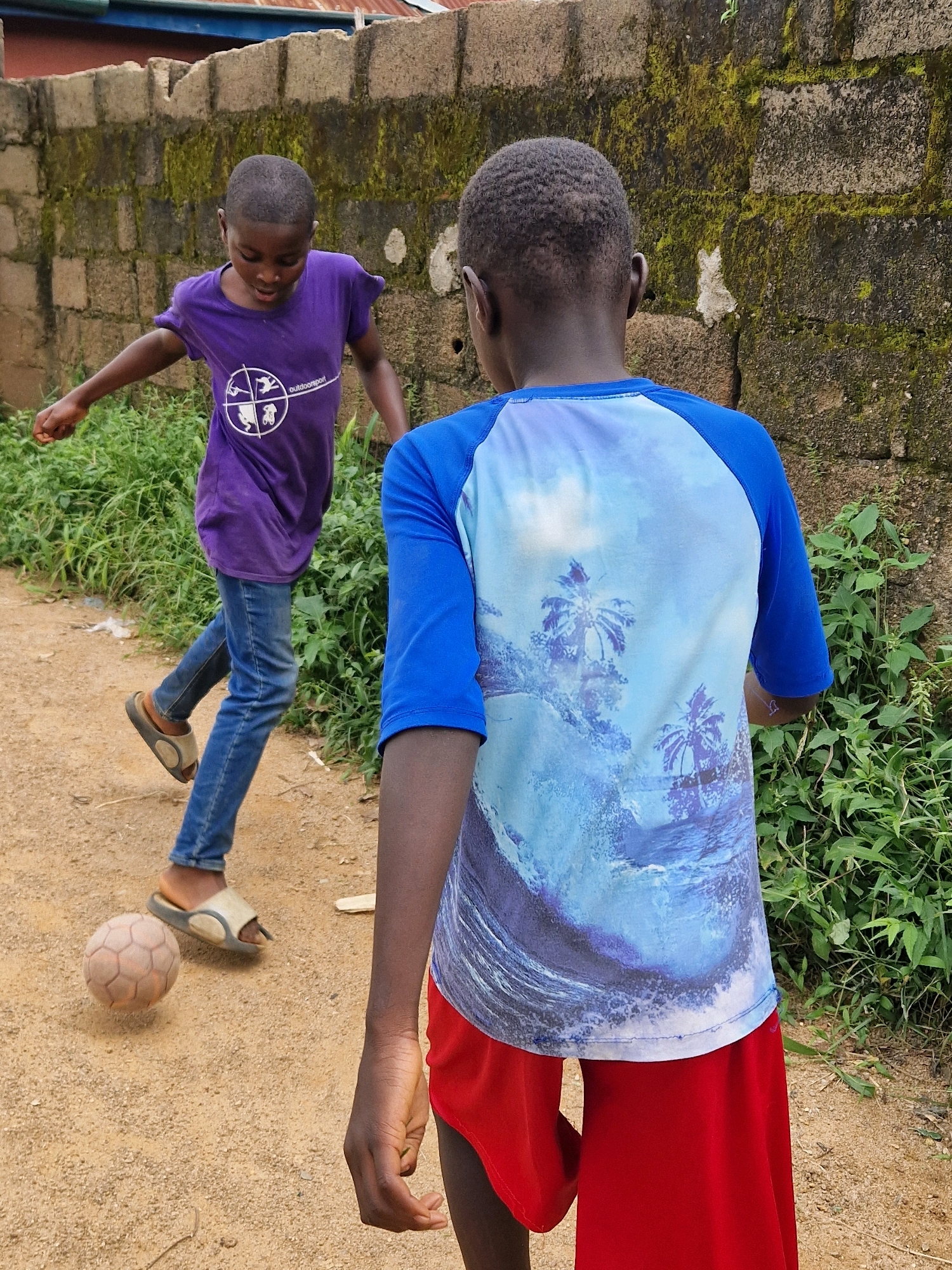 In an outdoor scene, two young boys are engaged in a game of soccer. The boy on the left, facing away from the camera, wears a purple t-shirt and blue jeans, and is in motion, kicking a soccer ball. The boy on the right, also with his back to the camera, is wearing a blue t-shirt with a tropical print and red shorts. The background consists of a textured, moss-covered brick wall and lush green vegetation. The ground is a light brown, dusty dirt path. The lighting suggests an overcast or daytime setting.