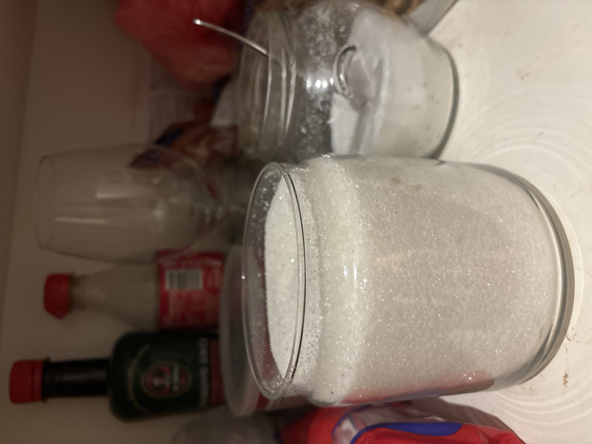 The image is a close-up shot of a shelf inside a cupboard. In the foreground, there are two glass jars filled with white granulated sugar. The jar on the left is larger and closer to the camera, filling most of the frame. The jar on the right is slightly behind and to the right of the first jar. A metal spoon is partially visible sticking out of the sugar in the right jar.
In the background, several other items are visible, though out of focus. These include a dark green bottle with a red cap on the far left, a clear bottle with a red cap behind the dark green bottle, and a clear wine glass. To the right of the wine glass, a red, somewhat amorphous object is visible, possibly a bag of something or a piece of fruit.
The shelf itself appears to be made of a light-colored material, possibly wood or laminate, and shows some signs of wear or smudges. The overall lighting is dim, suggesting the cupboard is not well-lit. The environment is domestic, likely a kitchen pantry or cupboard.
There are no people or animals present in the image, and no actions or interactions are depicted. The scene is static and focused on the arrangement of objects within the cupboard.
The location of Budva, Montenegro is not ascertainable from the image itself.