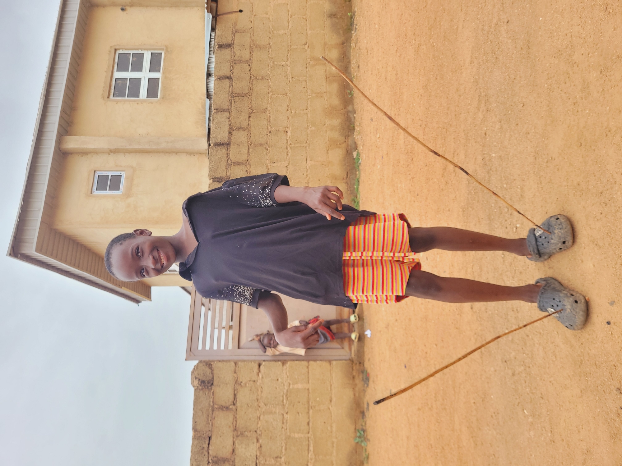 A young person is centered in the shot, facing the camera with a smile. They are wearing a dark top, striped shorts, and gray clogs. Two thin sticks, possibly used for playing or as tools, lay on the ground beside their feet. The background features a building with yellow walls and a white-framed window. To the side, there is a gate or fence constructed from light-colored bricks. The ground appears to be dirt or sand. Another younger child is barely visible behind the gate.
Although the location of the image cannot be confirmed from the image alone, the user prompt suggests the image was taken in Bukuru, Nigeria.