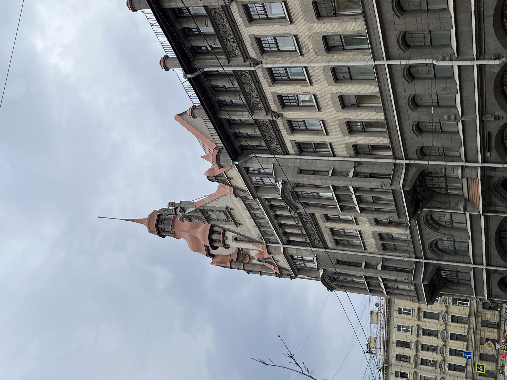 The image shows a tall building with a prominent spire in Saint Petersburg, Russia, shot from a low angle. The building's architecture is characterized by a mix of gray stone and brick with numerous windows arranged in regular rows. The spire, which is the most striking feature, has a reddish-pink hue at its base, contrasting with the gray of the main structure and the sky. The sky is overcast, and the presence of tree branches and utility lines suggests a street-level perspective. The overall impression is of a dense urban environment with historical architecture.