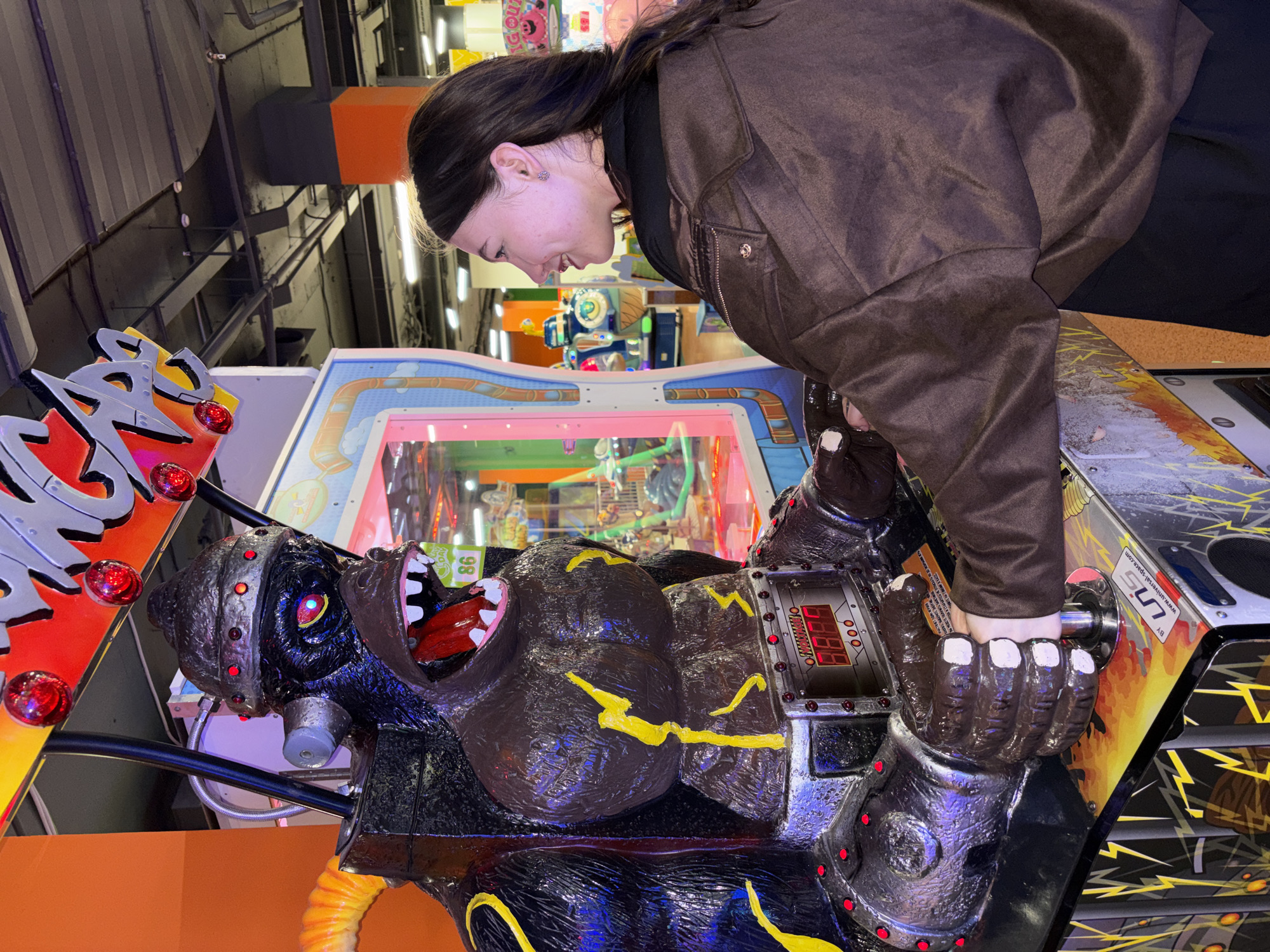 A woman is playing a gorilla arcade game. The woman is leaning towards the arcade game, pulling the handle of a robotic gorilla. She is wearing a brown jacket and a black top. The arcade game has a gorilla figure with an open mouth, and the game's console has a digital display. The environment seems to be an entertainment center or arcade. No information in the image confirms that this photo was taken in Perm, Russia.