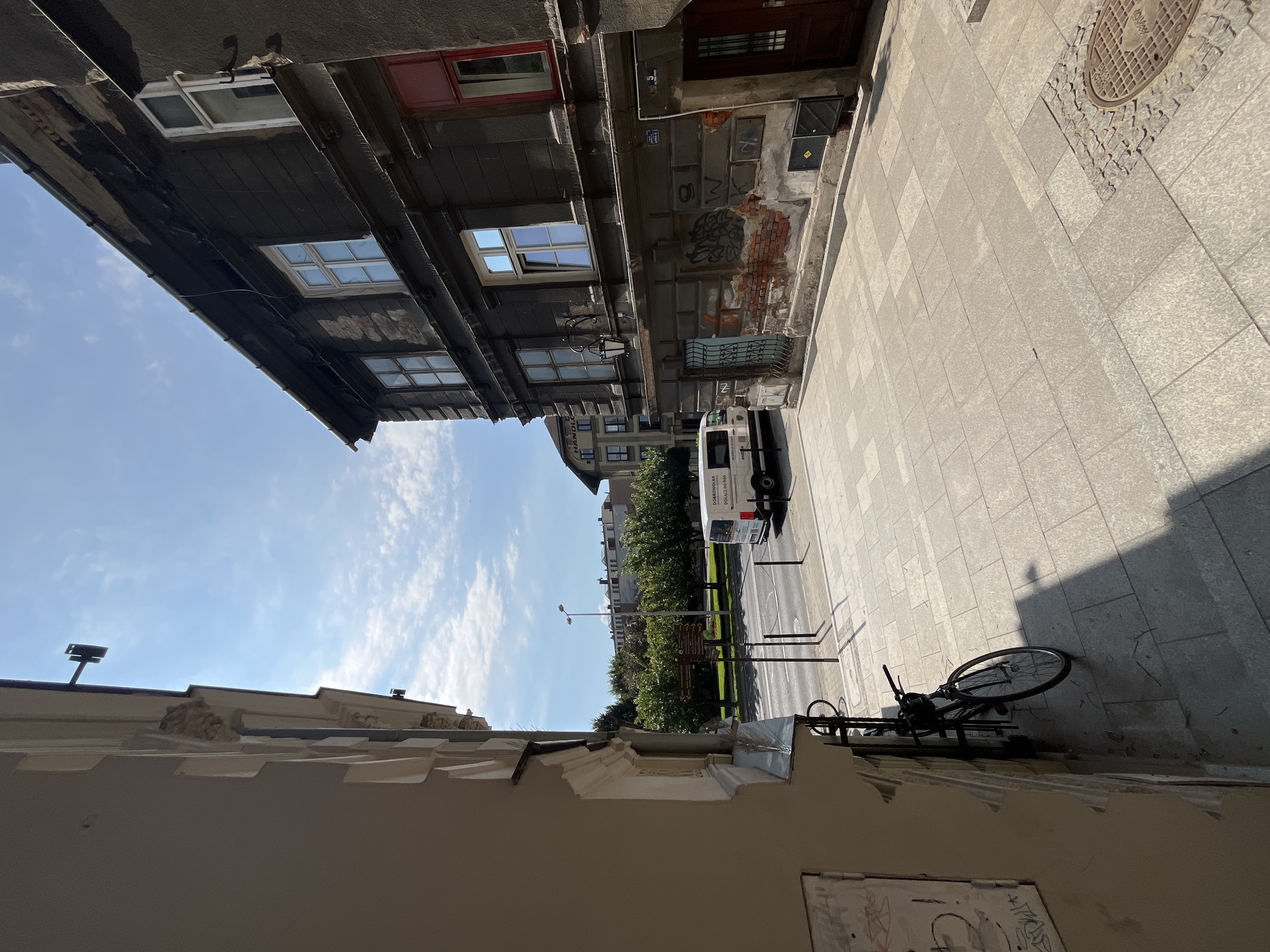 The image captures a street view in an urban environment under a bright, partly cloudy sky. On the right side of the frame, a dark, weathered building with multiple windows dominates the scene. Some windows are open, revealing reflections of the sky. The building's facade shows signs of age and disrepair, with exposed brickwork and graffiti. A red-framed window is visible on the upper floor, contrasting with the darker tones of the building.
To the left, a lighter-colored building runs parallel to the street. A bicycle is parked against its wall. The street in the foreground is paved with large, light-colored tiles, leading to a manhole cover. A white van with text on its side is parked on the street further down, alongside a tree-lined area and a road with traffic. In the background, more buildings are visible, suggesting a densely populated urban area. The overall impression is one of a historical urban setting on a sunny day.