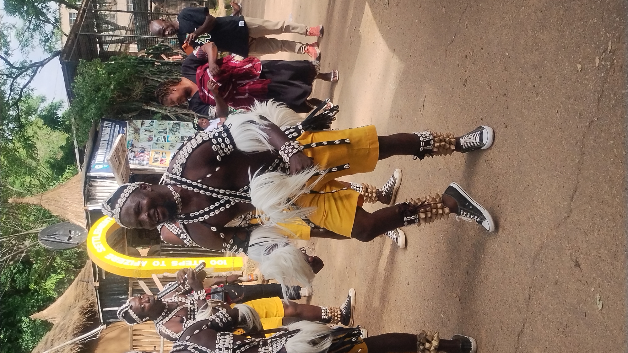 In an outdoor scene in Jos, Nigeria, a group of people are gathered. The focal point is a man in traditional attire, adorned with shells and white feather accents, performing a dance. He wears a beaded headdress, a beaded necklace and bracelets, and yellow shorts. His legs are decorated with shell and seed adornments above black sneakers. To his left, another man in similar attire plays a flute. Other individuals in traditional garb are visible in the background, as well as some spectators dressed in contemporary clothing. The environment features thatched-roof structures and a sign that reads "THINK TOURISM NIGERIA-TIN". The ground is paved with a dusty, light-colored surface.