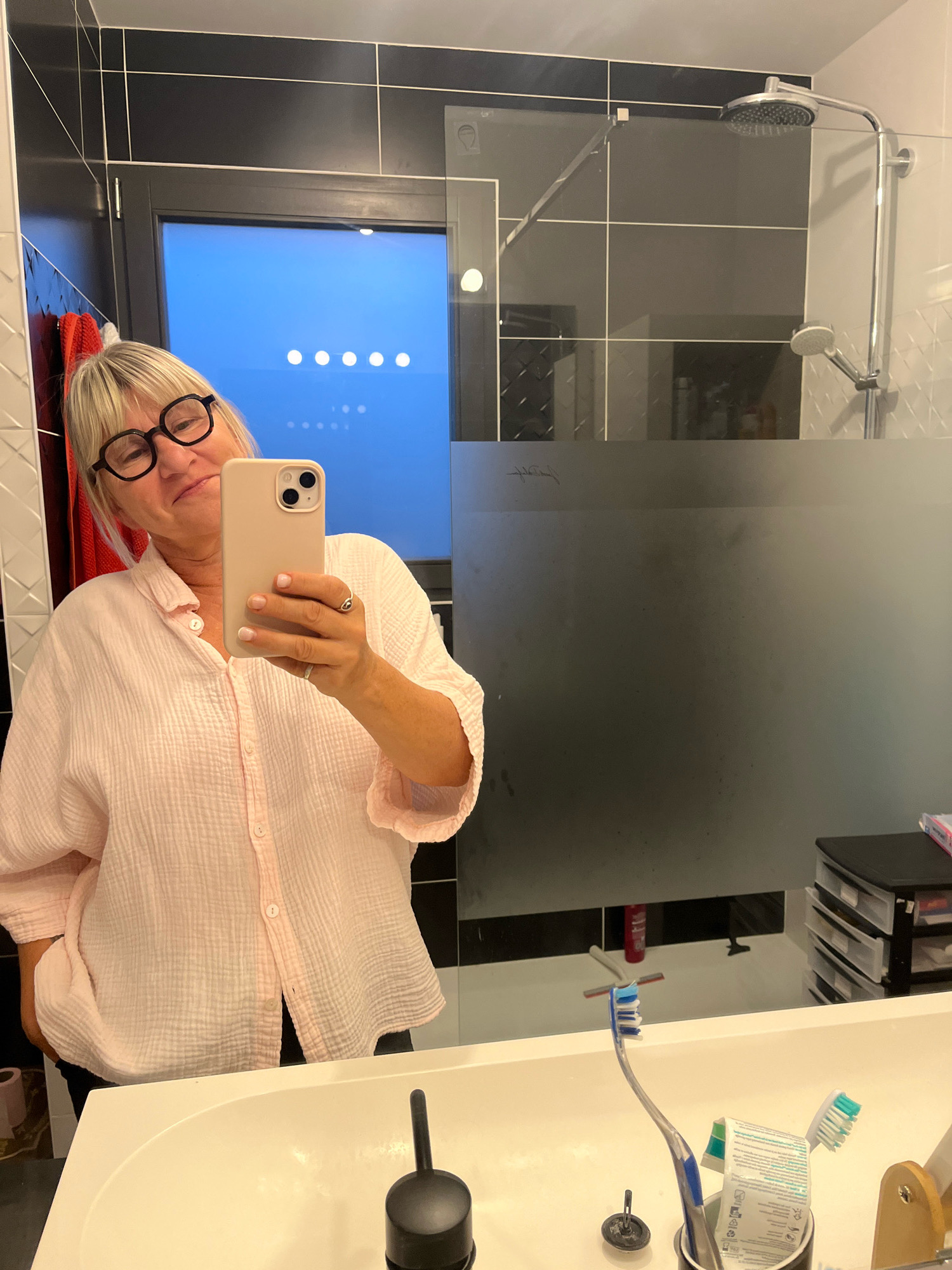 A middle-aged woman with blonde hair and dark-rimmed glasses is taking a mirror selfie in a modern bathroom. She is wearing a light pink, oversized button-down shirt. She is holding a light pink smartphone in her right hand, angled to capture her reflection. A ring is visible on her left hand. The bathroom features black and white tiled walls, a glass shower enclosure with a silver showerhead, and a white sink. On the counter, there are toothbrushes, toothpaste, and a black container with drawers. A window with blue light showing through is visible in the background. The overall impression is of a clean and organized personal space. The location is identified as Chambœuf, France.