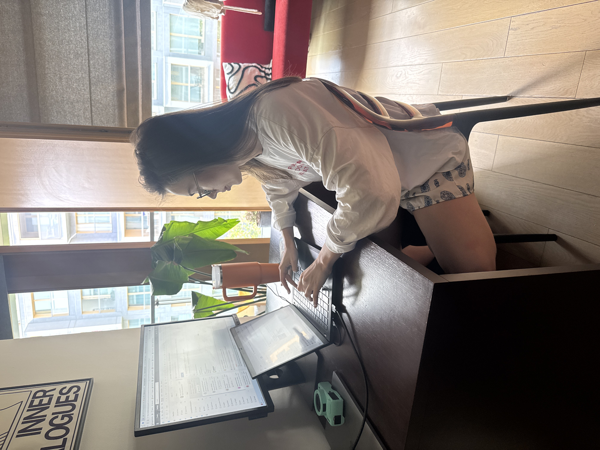 The image depicts a young woman intensely focused on her work at a home office setup, bathed in bright natural light, likely during the daytime.
**Subjects:** The central subject is a woman with long, dark hair and glasses, seen in profile, leaning forward over a desk. She is dressed in a long-sleeved white t-shirt and light-colored, patterned shorts. She is kneeling or crouching on a dark chair positioned behind the desk.
**Objects:** Her workspace features a dark wooden desk holding a laptop, on which she is actively typing. Two additional monitors are present: a larger upright screen and a smaller, portable one propped beside it. A light mint green device, possibly a portable speaker or camera, rests near the monitors. An orange insulated mug with a handle is positioned next to a large, vibrant green houseplant. In the background, a portion of a red sofa with a white patterned cushion is visible. The room has light-colored wooden flooring. Large windows reveal an exterior view of a modern building with numerous windows. A framed piece of wall art hangs on the wall to the left.
**Activity or event taking place:** The woman is actively engaged in typing on her laptop, likely utilizing the multiple screens for a work or study task. Her posture and gaze indicate deep concentration.
**Notable details:** The strong natural light through the windows suggests it is daytime with clear weather. The setting appears to be a modern indoor space, possibly an apartment or home office, given the residential furnishings and the view of other buildings. The woman's expression conveys focus and engagement. Visible text includes "INNER LOGUES" on the wall art and "FU**A DO WHA" (possibly "FUNNA DO WHAT" or "FUCKING DO WHATEVER") partially visible on her t-shirt due to a fabric fold.