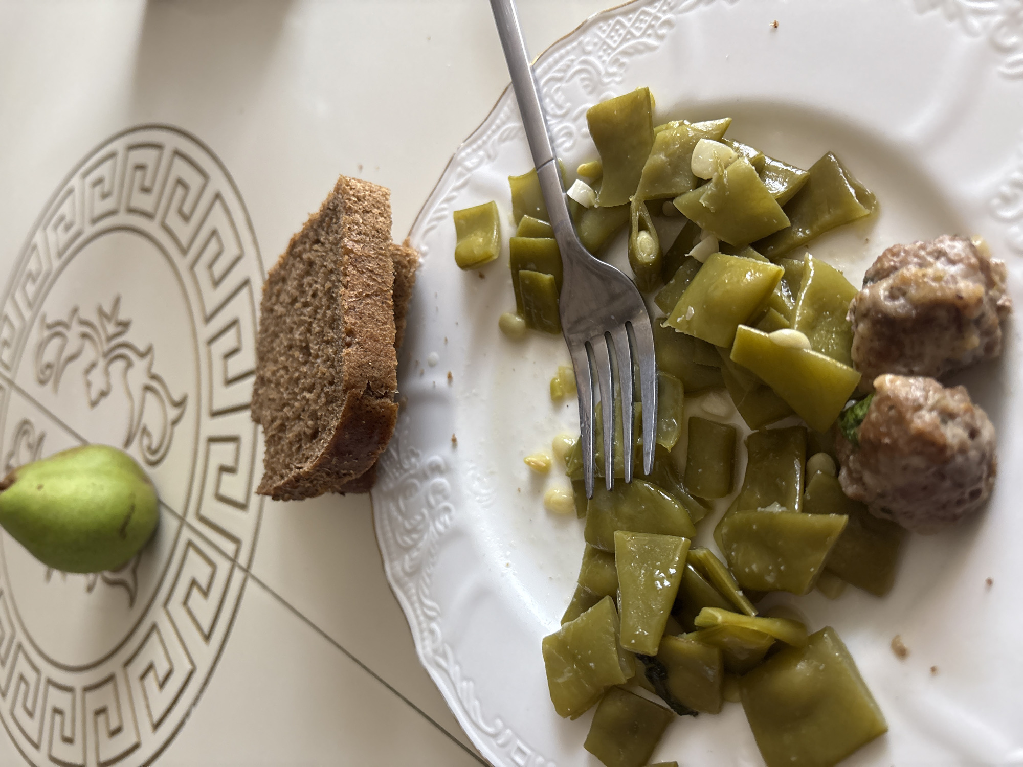 In Georgiyevsk, Russia, on a table with a patterned surface, a white plate holds a meal consisting of green beans, two meatballs, and a fork. Beside the plate, two slices of brown bread are placed, and in the background, a green pear rests on the table. The scene appears to be indoors.