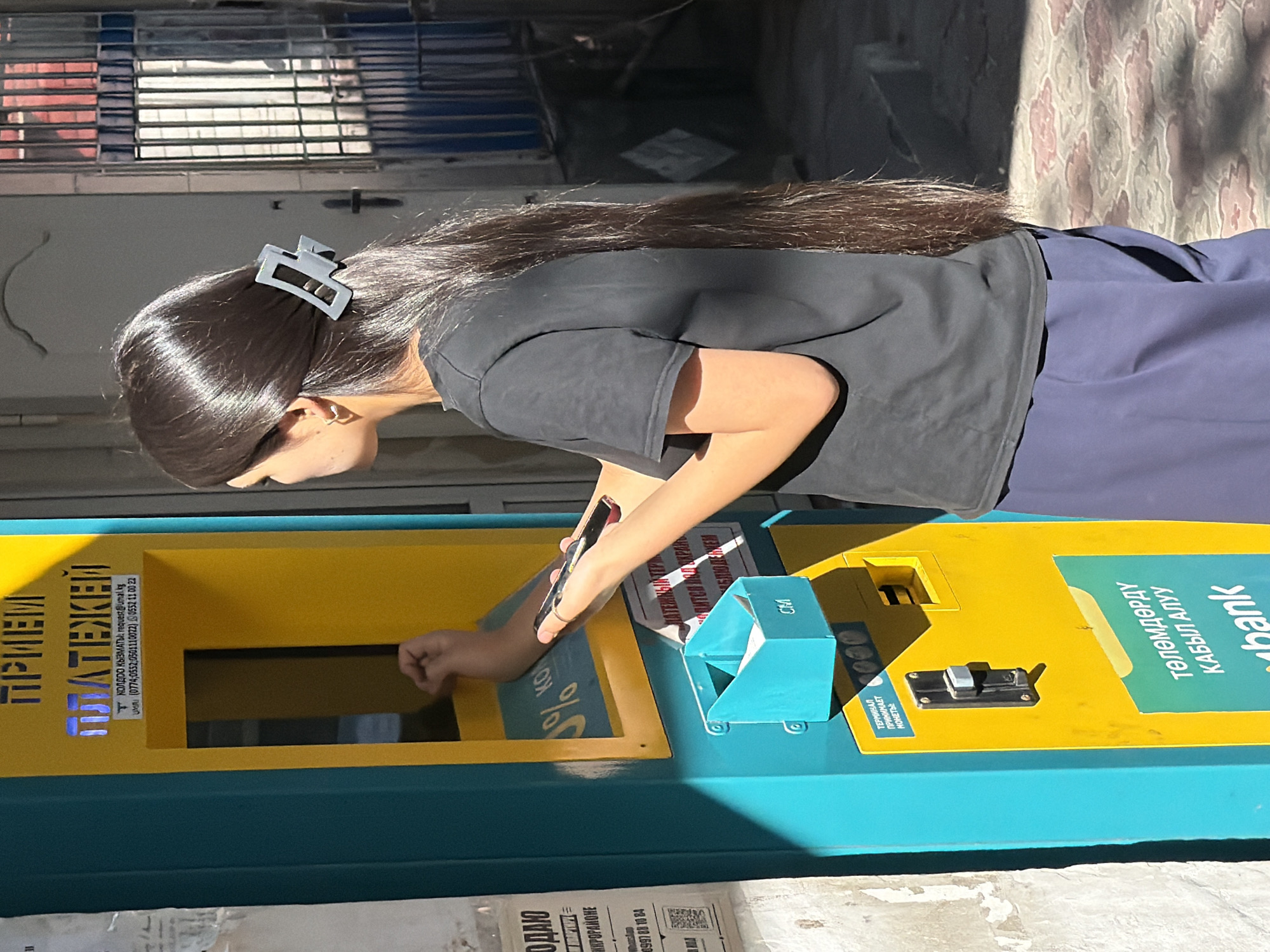 A young woman with long, dark hair, secured with a light gray hair clip, stands facing a bright yellow and turquoise payment terminal. She is wearing a black t-shirt and dark blue pants. Her right hand is extended towards the terminal's screen, holding a black smartphone in a red case. Her left hand is reaching into the terminal's slot, as if inserting something. The payment terminal has Cyrillic text and a small blue receptacle below a slot labeled "CM". The terminal is located outdoors on a paved surface with a patterned design. The lighting suggests it is daytime, with strong shadows cast by the terminal and surrounding structures. A partially obscured paper advertisement is visible on the wall to the left of the terminal.