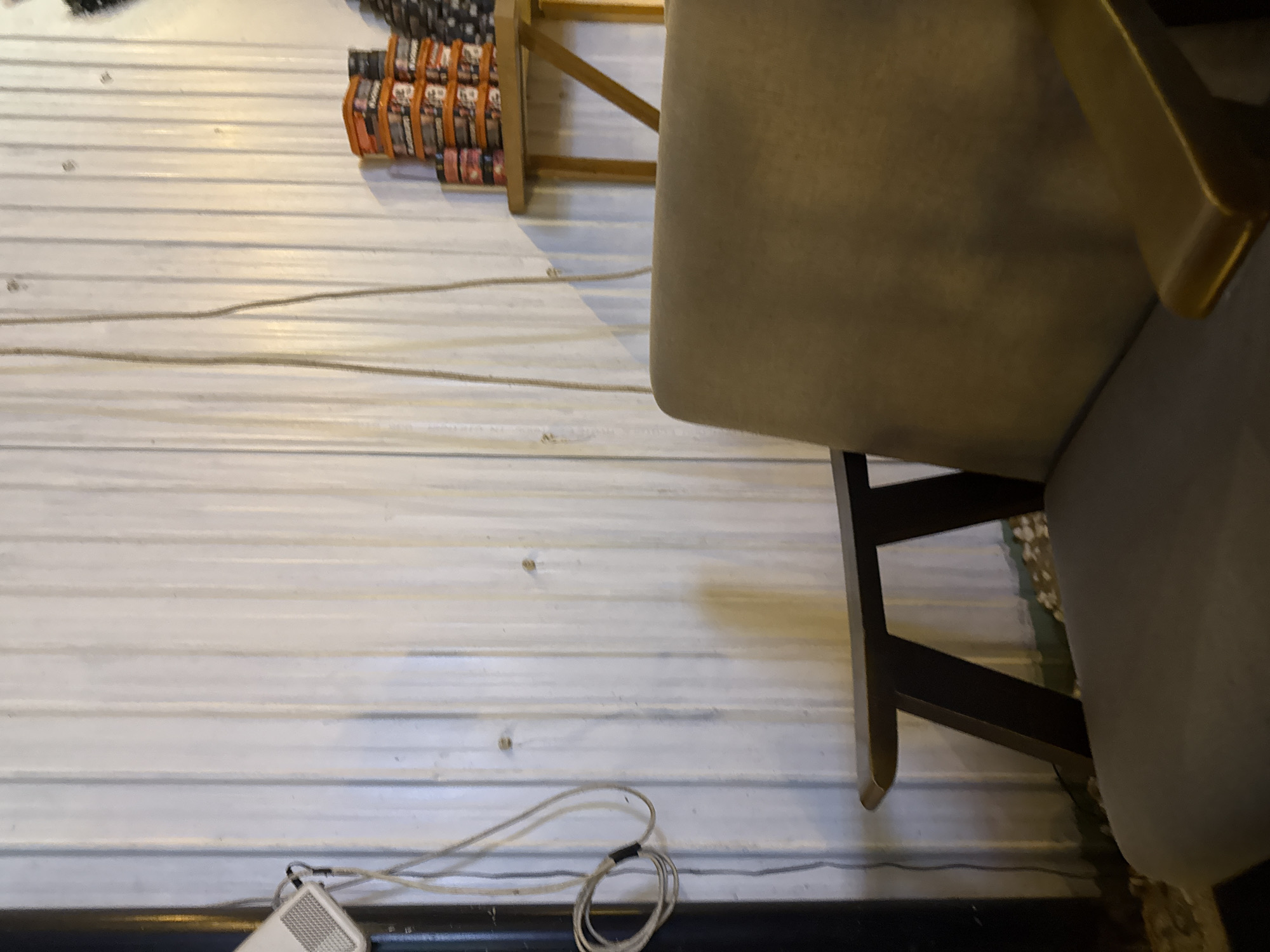 This image captures an indoor scene, likely in Đà Nẵng, Vietnam, with a functional and slightly industrial aesthetic. The main subjects include a light beige upholstered chair with dark wooden legs, positioned on the right side of the frame. To the upper left, a wooden shelf or rack displays multiple rows of packaged items, predominantly in orange wrappers. These packages appear to be instant noodles or similar snacks, with some details suggesting a common Vietnamese brand like "Hảo Hảo."
The walls and floor are covered with a distinctive white, horizontally ribbed material, possibly corrugated metal or paneling, creating a uniform backdrop for the setting. In the bottom left corner, a white electronic device, resembling a speaker or modem, sits on the floor, accompanied by a tangled white power cord and a coiled cable. Several other cables are visible running across the ribbed floor.
The lighting appears to be artificial and somewhat dim, casting soft shadows, which makes it difficult to determine the time of day. No people are visible, and no specific activity is taking place. The overall impression is that of a casual eatery, a waiting area, or a small shop with basic furnishings and supplies.