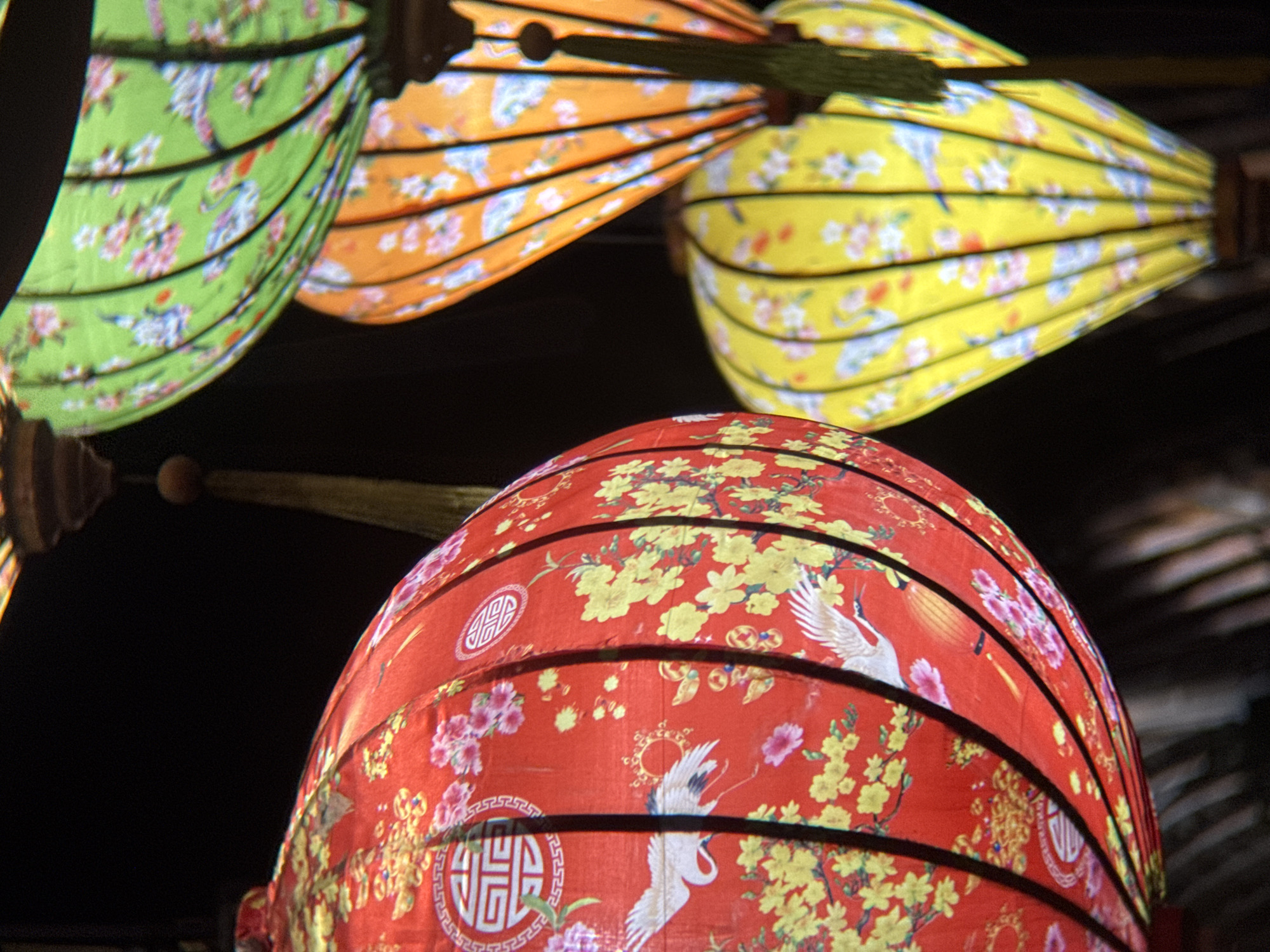 A close-up, low-angle shot captures a cluster of brightly colored, patterned lanterns hanging against a dark background. The lanterns are fabric and feature traditional floral and bird motifs. In the foreground, a large red lantern is adorned with yellow flowers and white cranes. To its right, a green lantern with similar patterns is partially visible, followed by an orange lantern, and a yellow lantern further back. The lanterns appear to be illuminated from within, creating a warm glow. The scene suggests an outdoor setting, possibly a night market or festival, in Nha Trang, Vietnam.