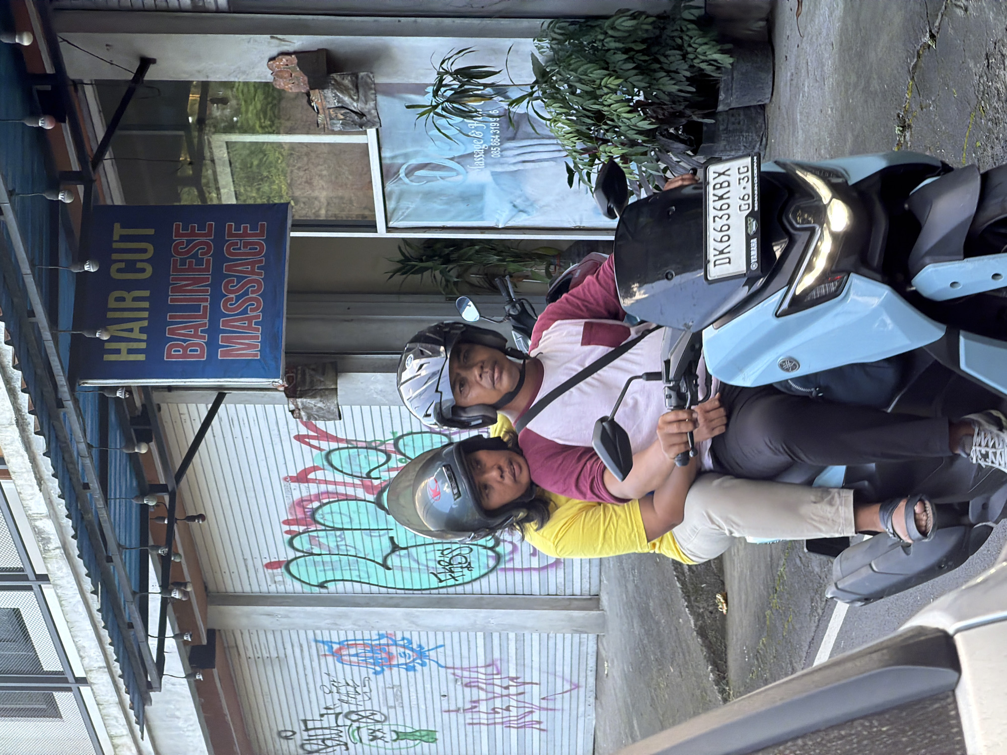 Two individuals, a male and a female, ride on a light blue scooter in front of a building in Ubud District, Indonesia. The male, wearing a maroon and white t-shirt and a black helmet, drives the scooter. The female, in a yellow t-shirt and light beige pants, is seated behind him, her arms around his waist. She also wears a black helmet. The scooter has a license plate that reads "DK 6636 KBX". Behind them, the building has a blue sign that reads "HAIR CUT BALINESE MASSAGE". The building's facade includes a corrugated metal roof and roll-up garage doors covered in graffiti. To the right of the sign, a glass door reveals an interior with potted plants and a poster with text and a phone number. The ground is paved with asphalt.