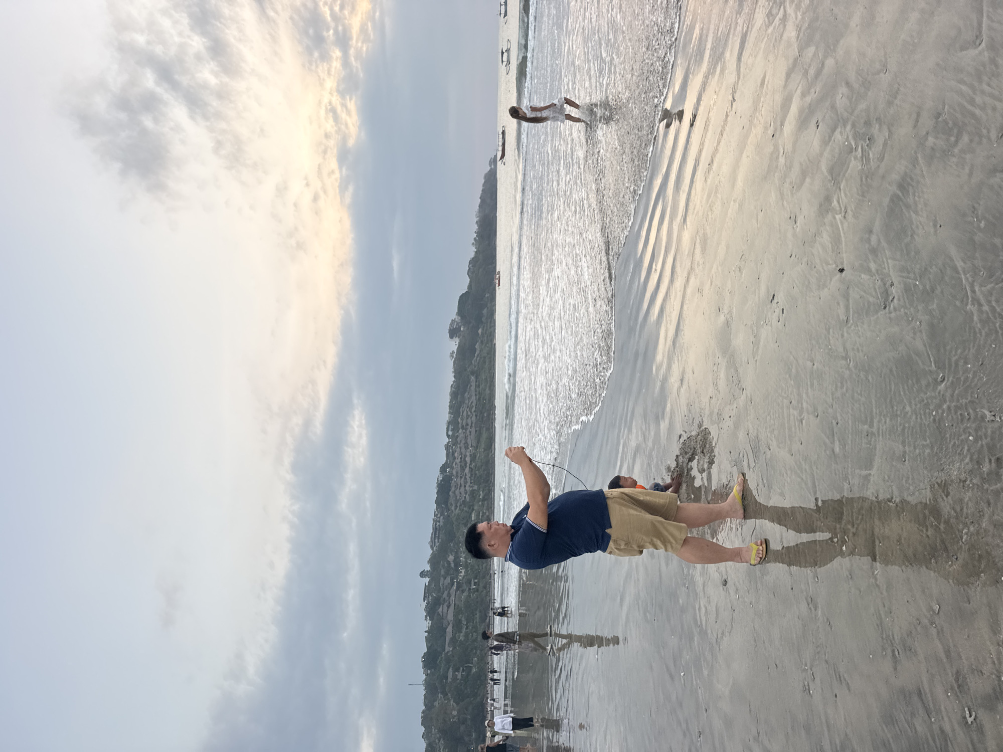 The image depicts a beach scene in Kuta Selatan, Indonesia, during what appears to be late afternoon or early evening. The sky is overcast with some patches of light filtering through, suggesting the sun is setting or has recently set. The ocean is calm with gentle waves lapping at the shore. The sand is wet and reflective, showing ripples from the receding water.
In the foreground, a man wearing a dark blue polo shirt and light brown shorts is standing and holding a smartphone, appearing to take a picture or video. He is wearing yellow flip-flops. To his right and slightly behind him, a young child is kneeling in the sand, seemingly playing.
Further down the beach, other people are visible walking and standing near the water. In the middle ground, a young woman in a white dress is walking through the shallow water, her reflection visible on the wet sand. Several small boats are anchored or moving on the sea in the background.
The coastline is lined with hills covered in buildings and vegetation, forming the backdrop of the scene. The overall atmosphere is relaxed and recreational, characteristic of a beach location.