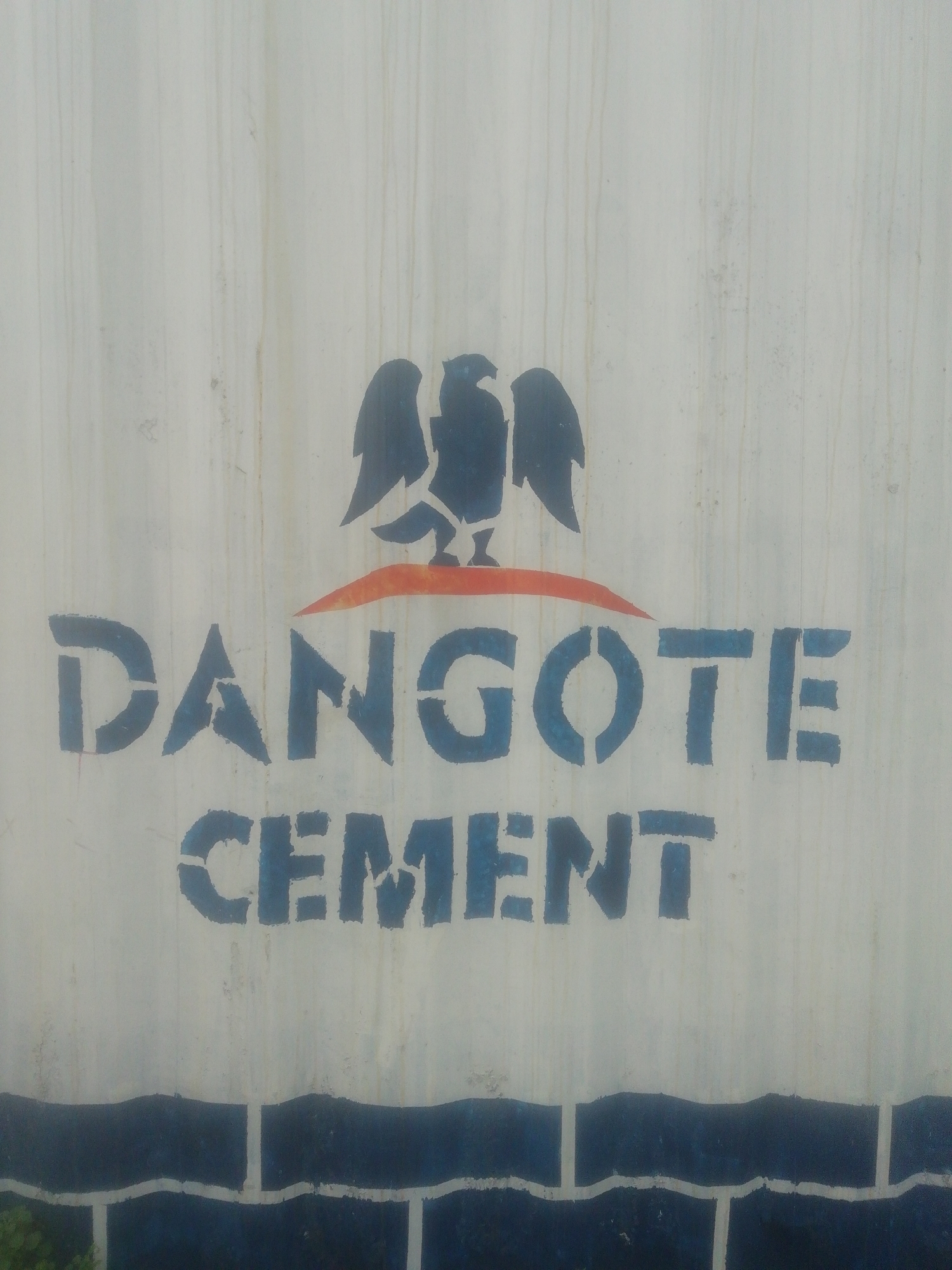 A close-up shot of a painted surface, possibly a wall or a container, displays the logo and name "DANGOTE CEMENT" in a stenciled, blue font. Above the name is an emblem featuring a bird-like figure, also in blue, positioned above an orange arc. The background is a light grey or white, and there are indications of wear and possible water stains. The bottom edge of the frame features a blue and white painted design, perhaps resembling a brick pattern. Vegetation is visible in the lower left corner of the frame. The location is unknown.