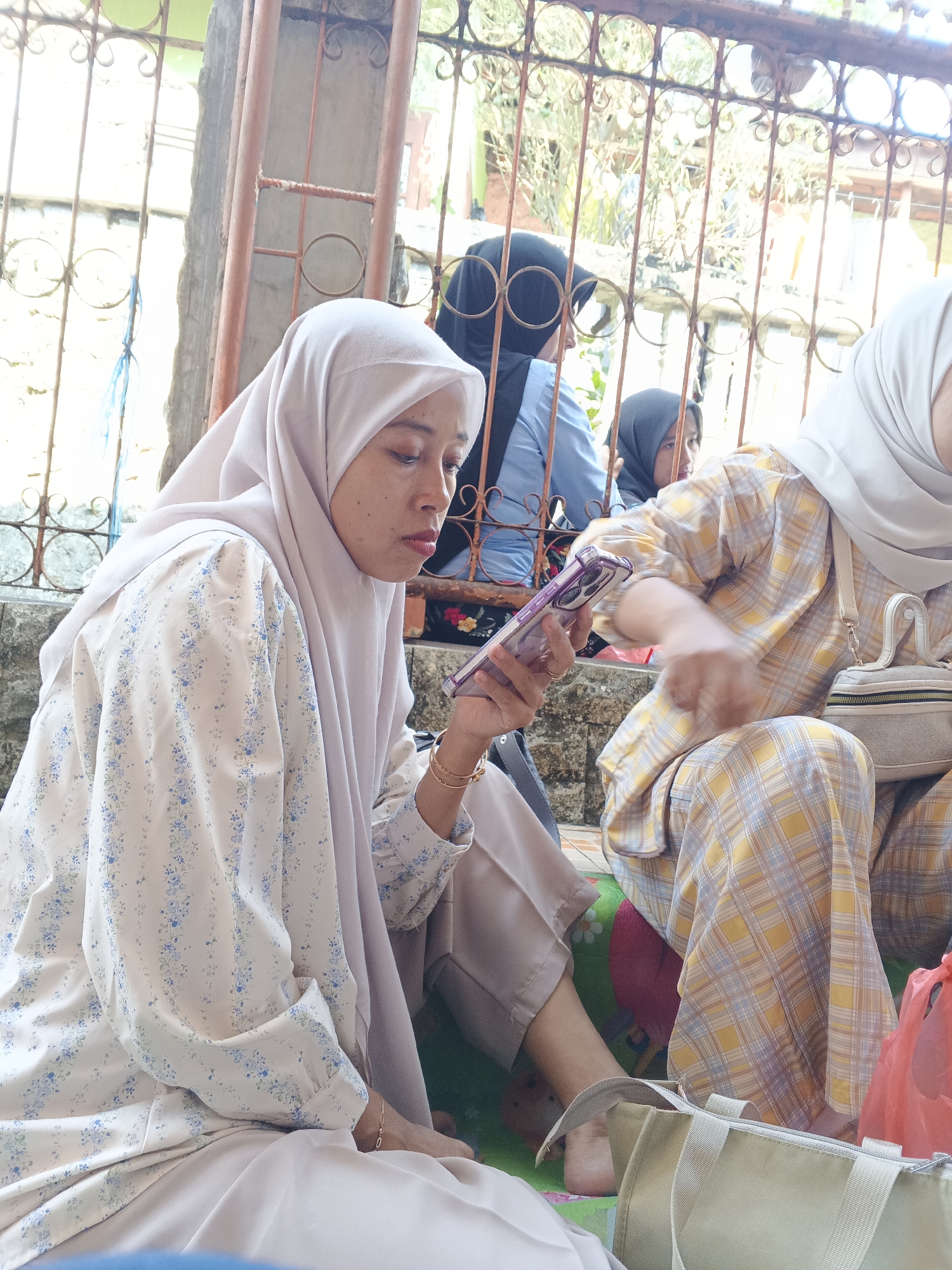 In Warungkiara, Indonesia, three women are seated outdoors near a metal fence. The woman in the foreground, wearing a light-colored hijab and a floral-patterned shirt, is looking down at a mobile phone in her hand. To her right, another woman in a plaid outfit is also seated, with a small bag on her lap. Further in the background, a third woman in a dark hijab is partially visible. The environment suggests a casual gathering or wait, with the women sitting on what appears to be a mat on the ground. The metal fence behind them has decorative scrollwork and provides a view of foliage and structures beyond.