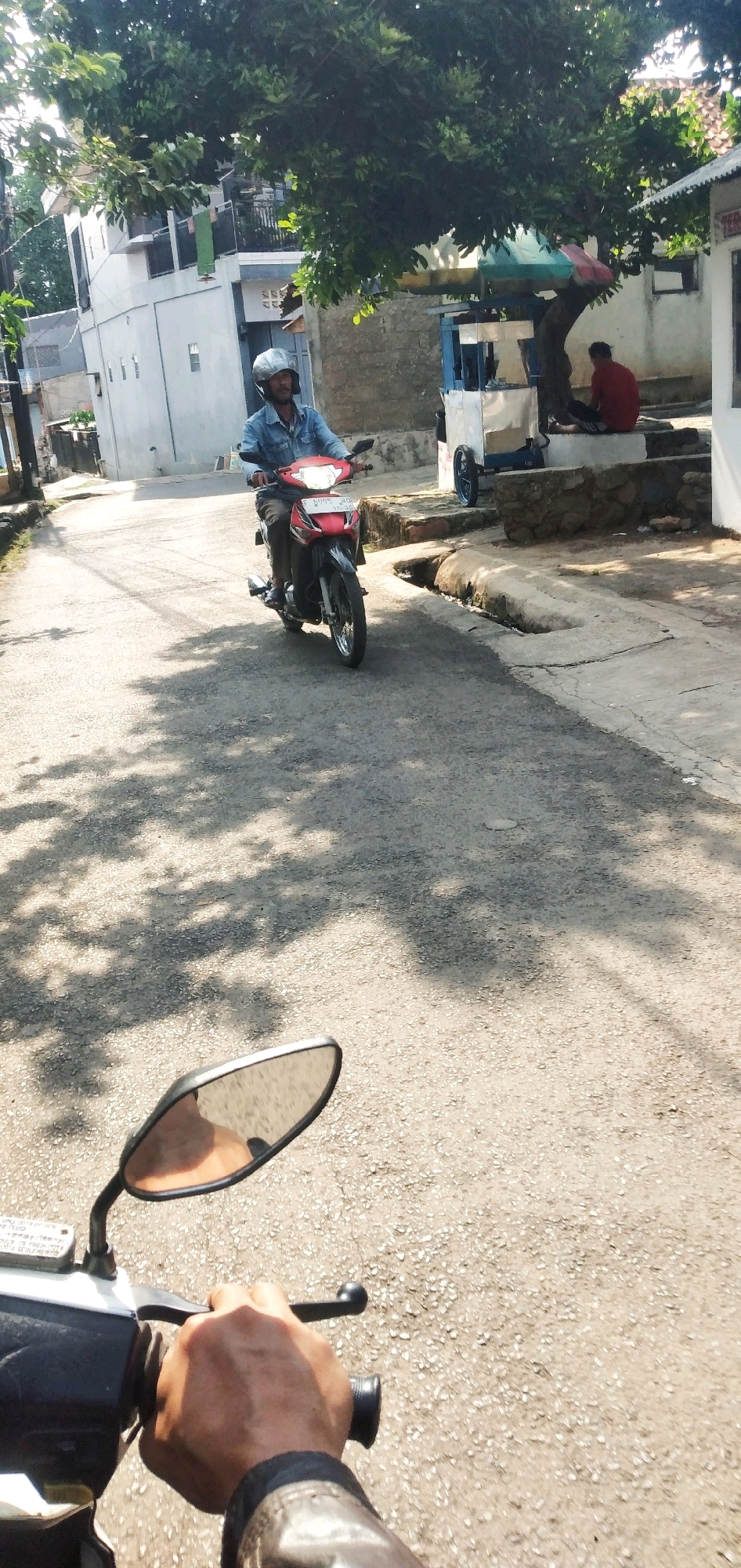 In a street in Cilaku, Indonesia, a man wearing a denim jacket and a helmet rides a red motorcycle. He is on a paved road lined with buildings and trees. The sunlight casts shadows on the road from the trees. In the background, a person in a red shirt sits under a tree near a small cart. The perspective of the photo is from another rider on a motorcycle, with a mirror and handlebar visible in the foreground.