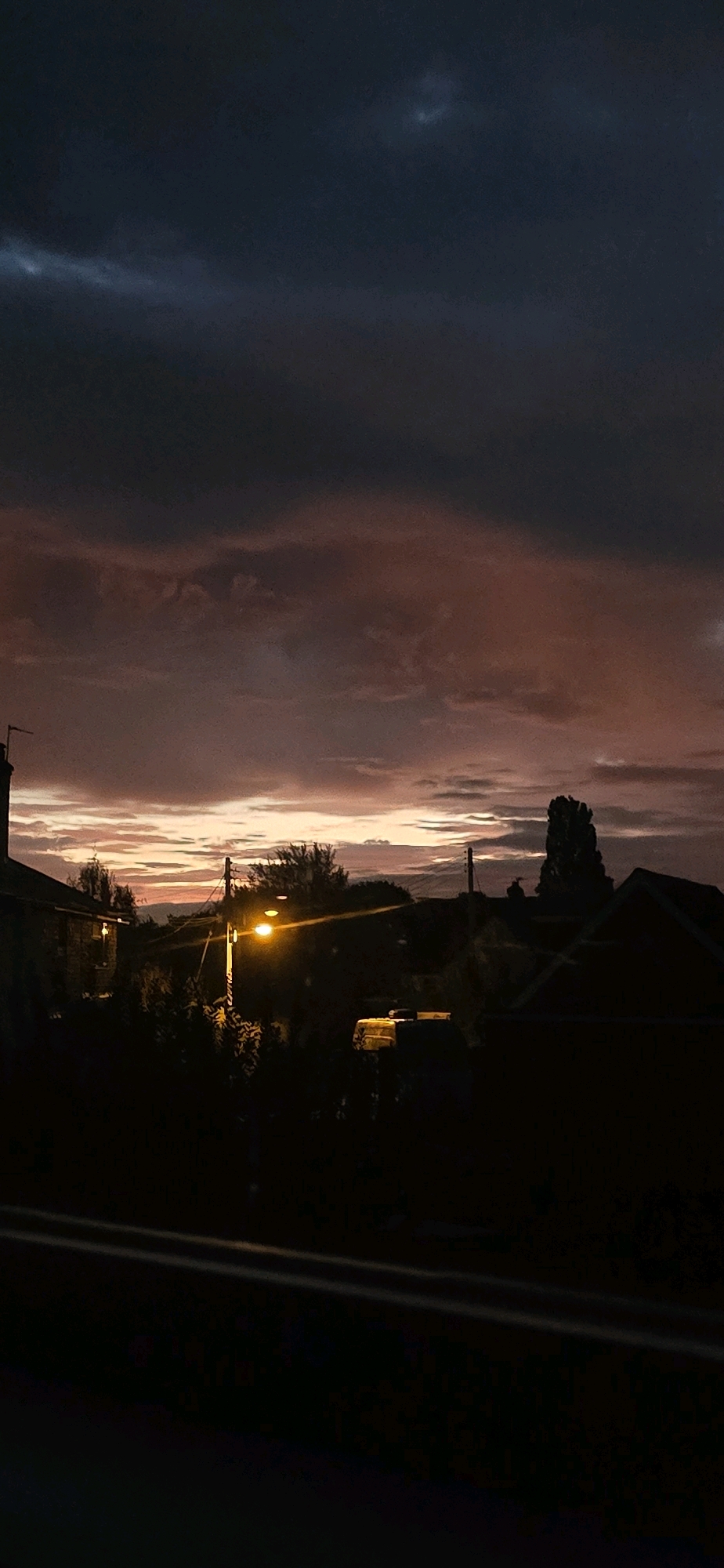 This is a vertical, low-light photograph taken at dusk or dawn, likely from an elevated position looking towards a suburban landscape in Bayston Hill, United Kingdom. The sky dominates the upper portion of the image, displaying a dramatic gradient of colors from deep indigo and dark gray clouds at the top, transitioning to hues of muted pink, orange, and pale yellow near the horizon. These colors suggest a sunset or sunrise with significant cloud cover.
Below the sky, the middle ground features a silhouetted row of buildings and trees. A prominent street lamp with a bright orange glow is visible on the right side of the image, illuminating a section of the surrounding foliage. To the left, the corner of a building with a lit window is partially visible. In the center, a few vehicles, appearing as dark shapes, are parked or positioned. The silhouette of a tall, slender tree is noticeable on the far right, against the colorful sky.
The foreground is almost entirely black, obscuring details, but it appears to be a dark surface, possibly a road or a railing. The overall impression is one of a quiet, residential evening scene in a rural or suburban setting. The lack of any visible people or specific activities suggests a captured moment of natural beauty and urban infrastructure at the transition between day and night.