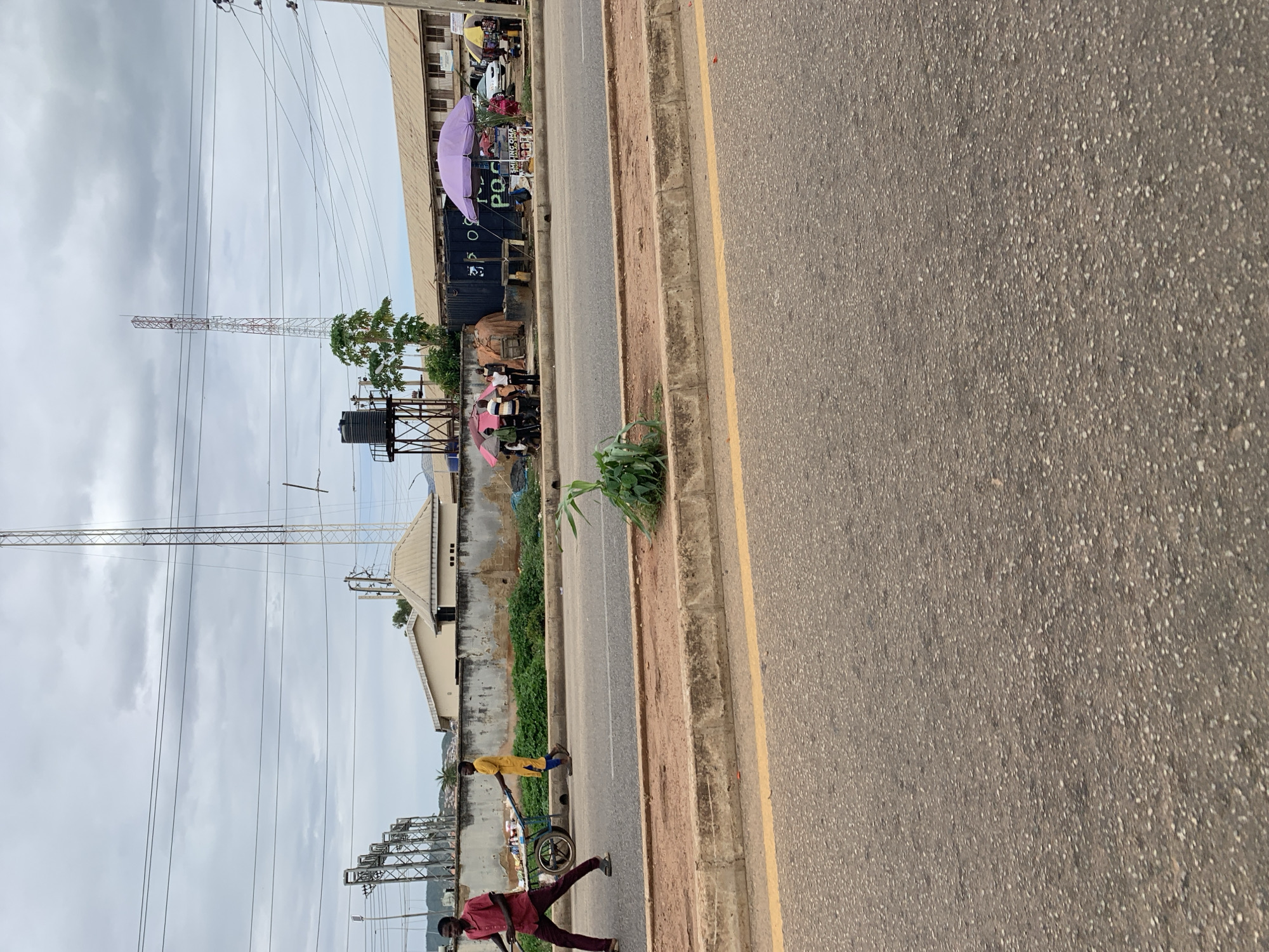 This image captures an outdoor street scene in New Stadium, Nigeria, under a heavily overcast sky. The foreground features a paved asphalt road with a faint yellow line near the curb. A raised dirt and gravel median strip separates the road from a pedestrian area, where a small green plant grows.
Along the mid-ground, behind the median, a low, weathered concrete wall extends horizontally. Two male individuals are present near this wall, moving from left to right. The first individual, wearing a red shirt and dark trousers, pushes a blue two-wheeled cart carrying various items. The second individual, dressed in a yellow tunic and dark trousers, walks behind or alongside the cart.
Beyond the wall and to the right, an informal market area is established. A dark, container-like structure, partially obscured, displays white lettering including "P.O.C". Approximately five to seven individuals are gathered in this area, sheltered by multiple open umbrellas of varying colors, including a prominent large purple umbrella, and smaller ones in red/white stripes, green, and yellow. A sign visible beneath the purple umbrella reads "SMILING OMA SALAD SPOT".
The background displays a complex urban landscape. On the left, a series of metal lattice utility structures and multiple power lines are visible. Further to the right, a black water tank rests on a metal stand. Several trees, including papaya and palm trees, are scattered throughout the scene. Two tall telecommunication towers stand prominently against the sky: a taller white/grey lattice tower and a shorter red/white lattice tower. Various buildings, including a light-colored structure with a ridged roof and a longer, multi-story building with numerous windows, are visible in the background. Distant hills or elevated terrain are discernible on the horizon. The sky is uniformly grey with dense cloud cover.