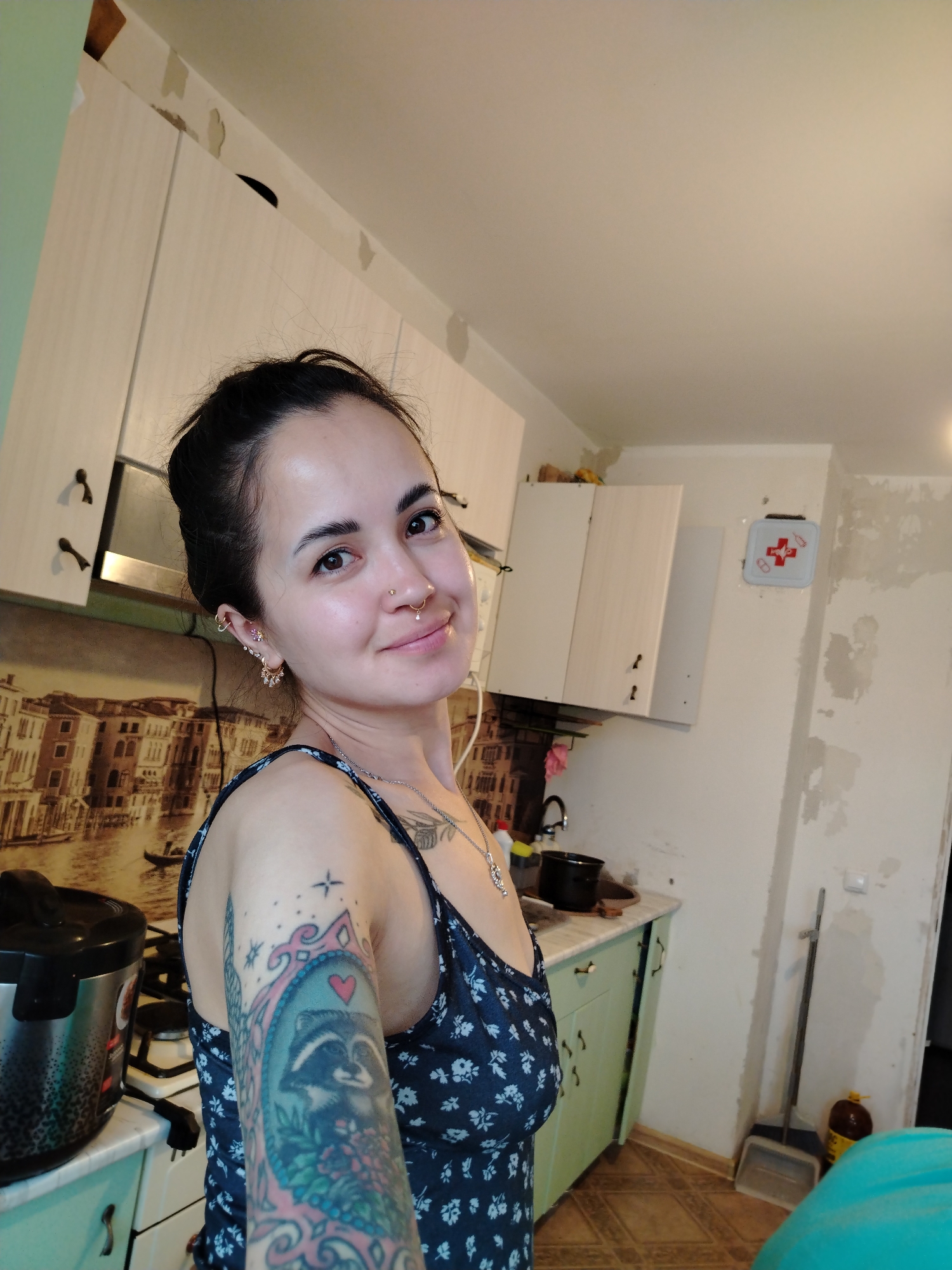 A young woman with dark hair in a bun, wearing a dark blue floral print dress, poses for a selfie in a kitchen. She has multiple piercings in her ears and nose, and a large tattoo of a raccoon surrounded by flowers on her left arm. The kitchen appears to be undergoing renovation, with unfinished walls and exposed plaster. Cabinets are visible, as well as a stove, a pot, and a first aid kit mounted on the wall. The background features a mural depicting a Venetian cityscape. The location is identified as Noviy, Russia.