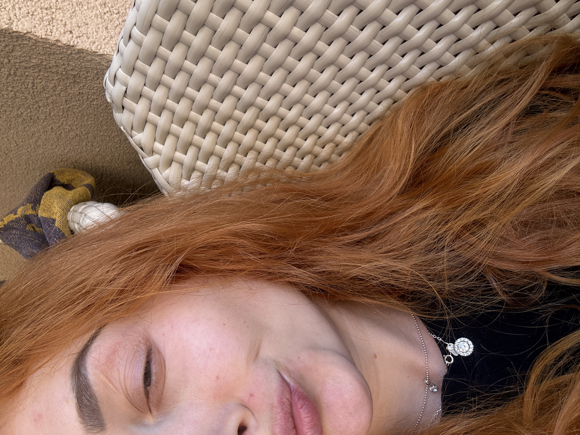 A top-down, close-up shot features a woman lying on a wicker chaise lounge, likely outdoors. The location is possibly in Budva, Montenegro, based on the prevalent outdoor lifestyle and architectural styles common in Mediterranean coastal towns. The woman has fair skin and red hair which is spread across the top portion of the lounge. She is wearing a silver necklace with a circular pendant. Part of a yellow and gray fabric item is visible at the edge of the lounge. The background consists primarily of the woven texture of the lounge's headrest and cushions.