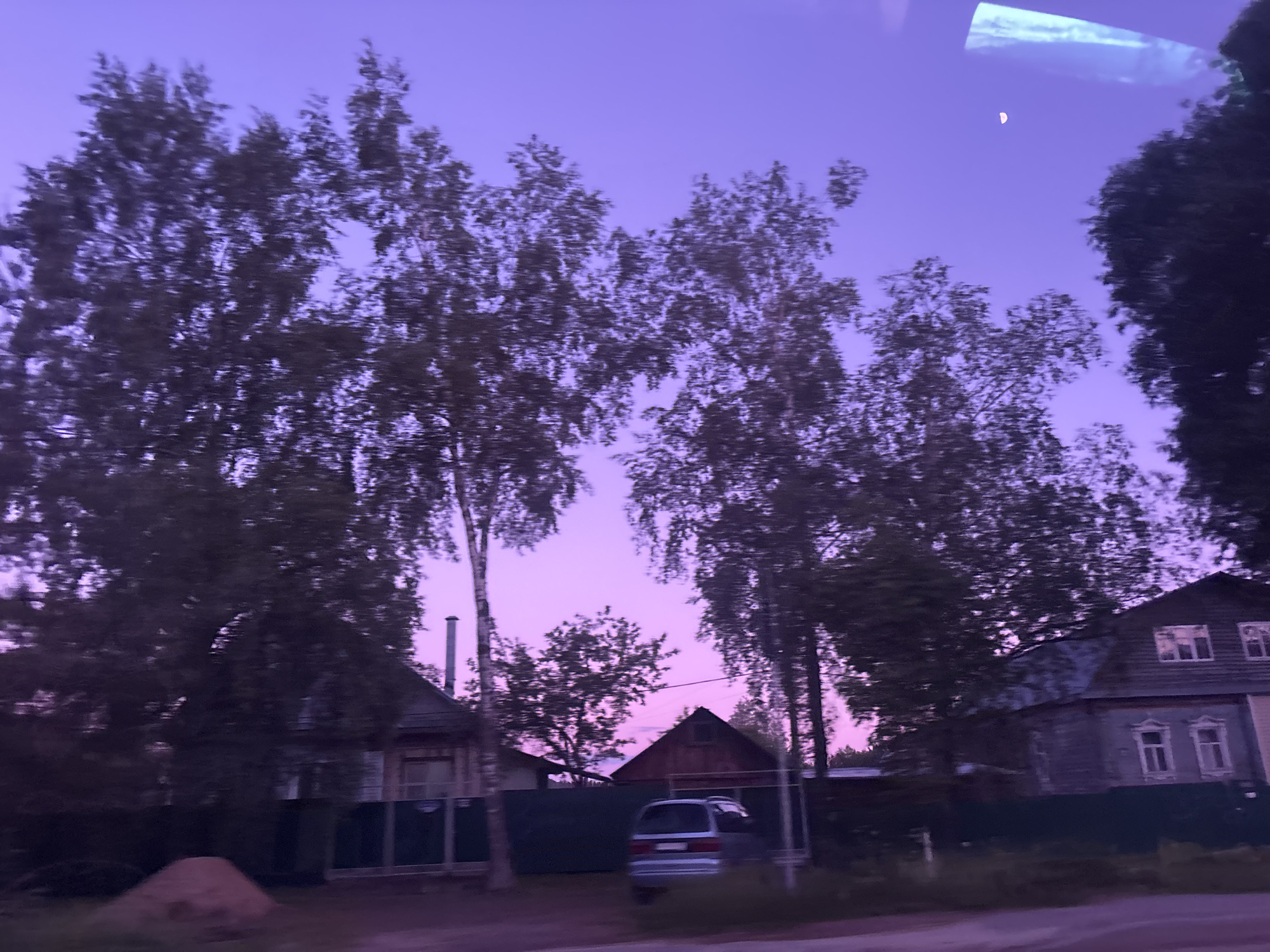 The image depicts a twilight or dawn scene in the Zaprudnya urban settlement of Russia. Several trees with sparse foliage frame a view of houses and a silver car parked along the side of the road. The sky is a pale purple hue, possibly indicative of dusk or dawn. A sliver of the moon is visible in the upper right of the frame. The houses are of a traditional design, some appearing to be wooden structures, with visible windows and roofing details. A fence runs along the property lines. The overall atmosphere is quiet and still, with a blurred effect possibly due to movement. The scene captures a residential area in the Zaprudnya settlement.