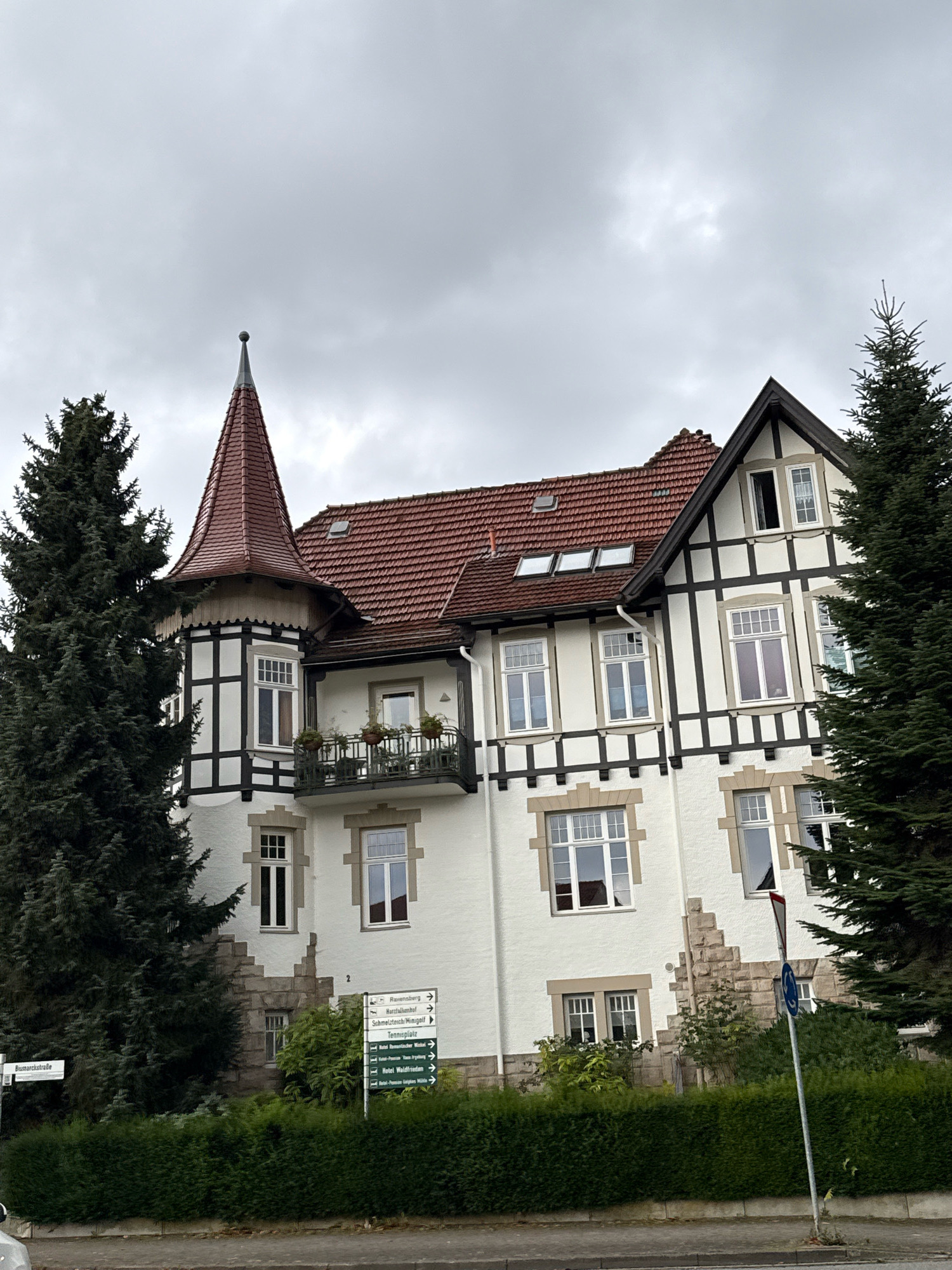 The image shows a building in Bad Sachsa, Germany, with a prominent tower and half-timbered architectural elements. The building is painted white with dark brown timbers and a red tiled roof. It features several windows, a balcony with potted plants, and dormer windows on the roof. Two large evergreen trees flank the building, and a neatly trimmed hedge runs along the front. A street sign reading "Bismarckstraße" is visible on the left, and a directional sign with multiple destinations, including hotels and tourist attractions, is located on the right. The sky is overcast with gray clouds, suggesting a cloudy day. There are no people visible in the image. The scene depicts a residential or possibly a hotel building in a green, tree-lined area.