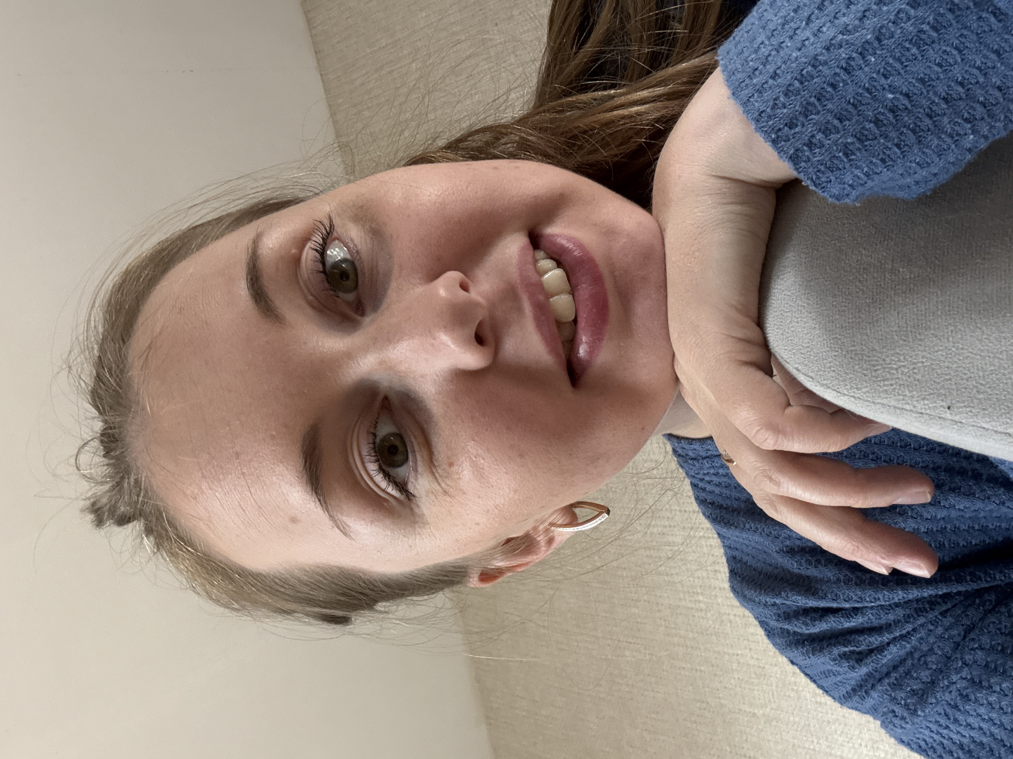 A woman is captured in a close-up, selfie-style photograph. She has fair skin, light brown hair pulled back from her face with some strands loose, and green eyes. She is wearing a blue, textured sweater and a thin, gold hoop earring in her left ear. Her left arm is bent, and her hand rests on her chin, supporting her head. She is smiling slightly, with her lips parted to reveal her teeth. The background appears to be a textured, light-colored wall, suggesting an indoor setting. There are no discernible city landmarks or environmental cues to indicate a specific urban location.
