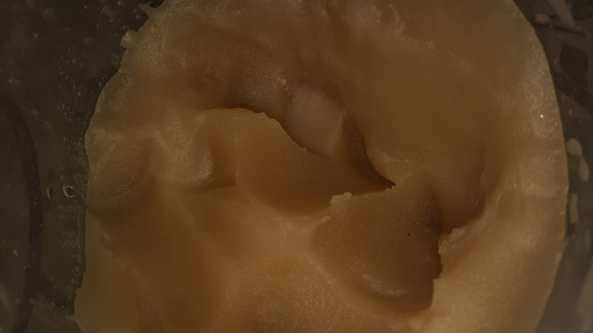 The image is a close-up, overhead shot of a thick, light brown substance, possibly a food item like mashed potatoes or dough, filling most of the frame. The texture is uneven and lumpy, with smooth, undulating surfaces and some rougher edges. There are subtle variations in color, ranging from pale yellow to a slightly darker tan. The substance appears to be contained within a transparent vessel, the side of which is visible on the right and left edges of the image, showing a dark, speckled background and some small bubbles or water droplets. The lighting is dim, casting soft shadows and highlighting the texture of the substance. There are no people or clear actions depicted. The context of Шклоў, Belarus is not directly visible in the image itself.