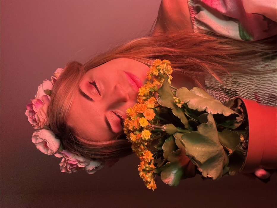 A woman lies with her eyes closed, face bathed in a reddish hue. She wears a flower crown of pink and white blooms. Beside her face, a potted Kalanchoe plant bursts with small, vibrant yellow-orange flowers and broad green leaves. Her hair is long and blonde. Her lips are pink. The scene is dreamlike, the colors saturated and soft. She appears serene and still.