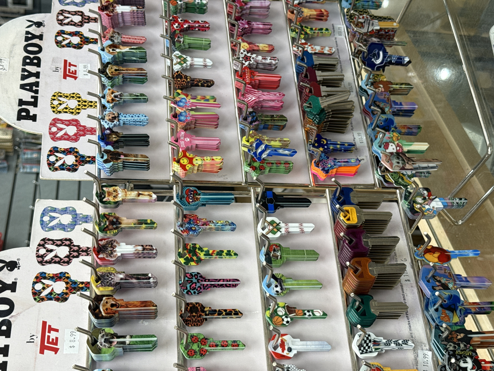 A collection of decorative keys displayed on a metal rack. The keys vary in design, including images of Playboy logos, animals, sports themes, cartoon characters, and abstract patterns. Signage on the rack advertises the keys are "by JET" and "PLAYBOY" with varying price points ($8.99 and $10.99). The context is retail. The image's location, the District of North Vancouver, Canada, cannot be determined from the image.