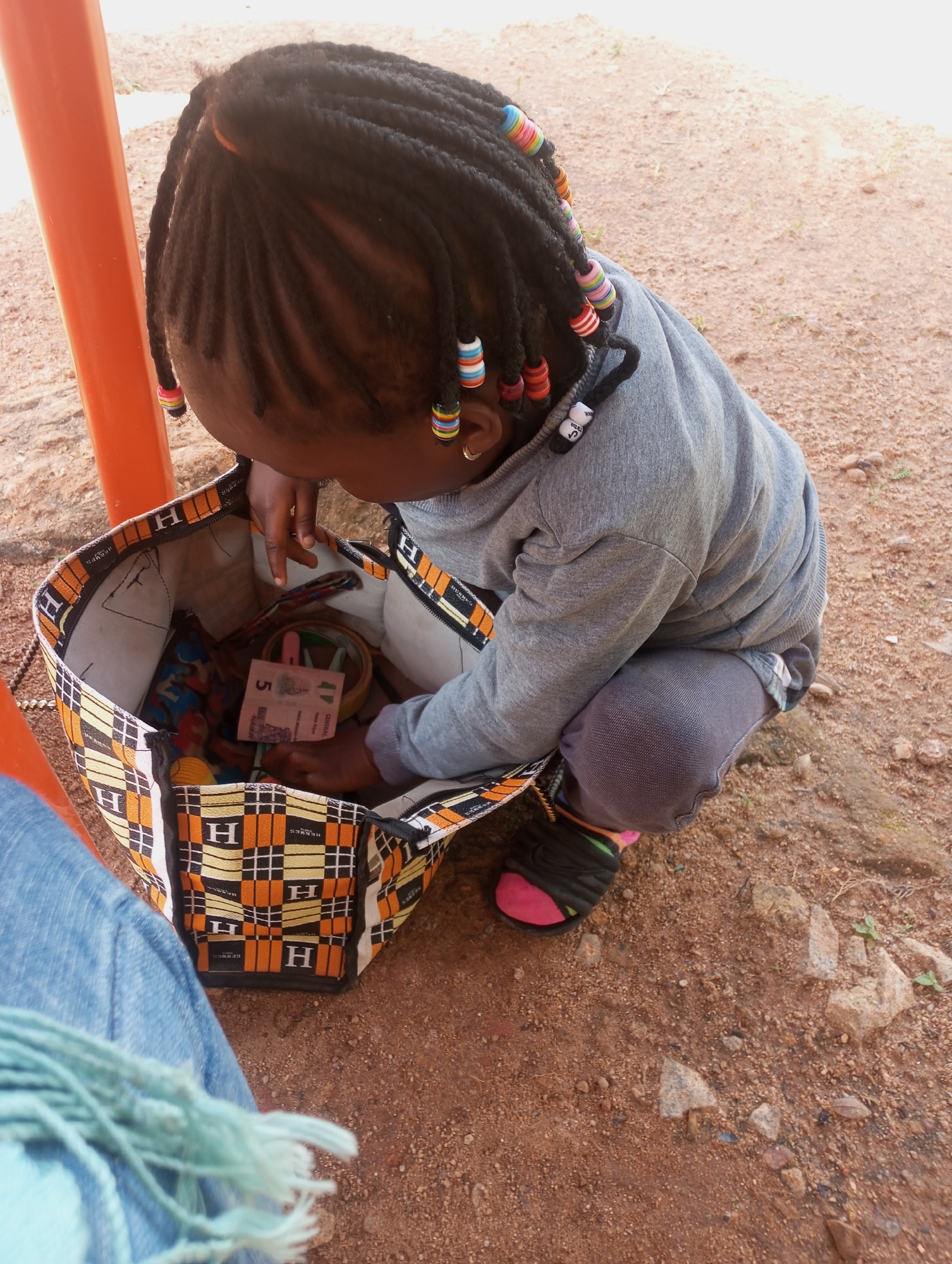 In the city of Jos, Nigeria, a young girl with braided hair adorned with colorful beads kneels on the dusty ground. She is looking into an open, patterned tote bag filled with various items, including what appears to be a 5-unit banknote. The girl is wearing a gray hoodie and dark pants, with one sandal visible on her right foot, featuring a pink accent. Her left leg is bent as she reaches into the bag. The surrounding environment is characterized by dry, reddish-brown earth scattered with small stones and sparse vegetation. An orange pole is partially visible in the background, suggesting an outdoor setting, possibly near a playground or public area.