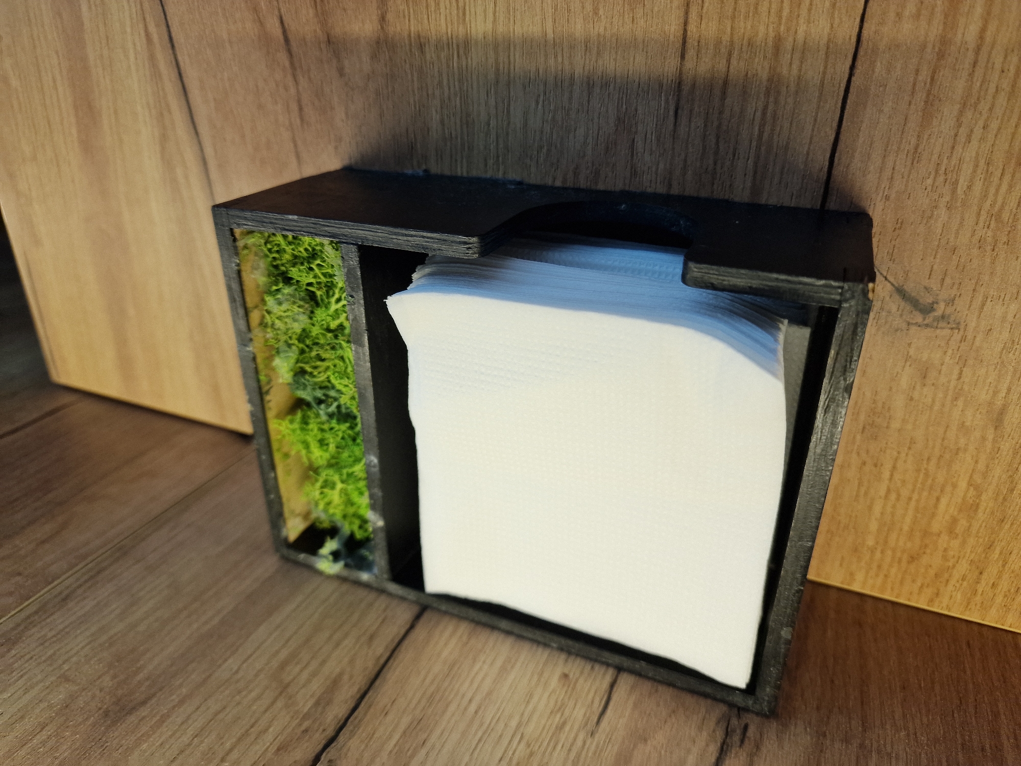 A black napkin holder, filled with white napkins, sits on a wooden surface. The napkin holder has a rectangular compartment at the top filled with green moss. The wooden surface consists of planks with visible grain.