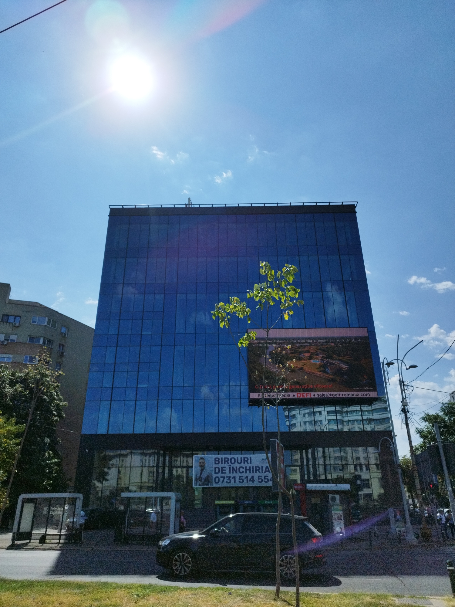 The image shows a modern glass-fronted office building with a bright blue facade, reflecting the sky and clouds. A large digital billboard is mounted on the building, displaying an aerial view of a developed area with water features and greenery, along with text that includes "BIROURI DE ÎNCHIRIERE" (Offices for Rent) and a phone number, "0731 514 554". The building is located in Bucharest, Romania, as suggested by the presence of "Primăria Capitalei" (City Hall) on a bus stop shelter.
In the foreground, a dark-colored car is parked on the street. The street has a sidewalk with bus stops and trees. The sky is clear and bright with the sun prominently visible, causing lens flare. Other buildings are visible in the background, including a multi-story residential building to the left. Utility poles and wires are also present along the street. Several people are visible near the bus stops and on the sidewalk. The overall scene depicts a typical urban environment in Bucharest on a sunny day.