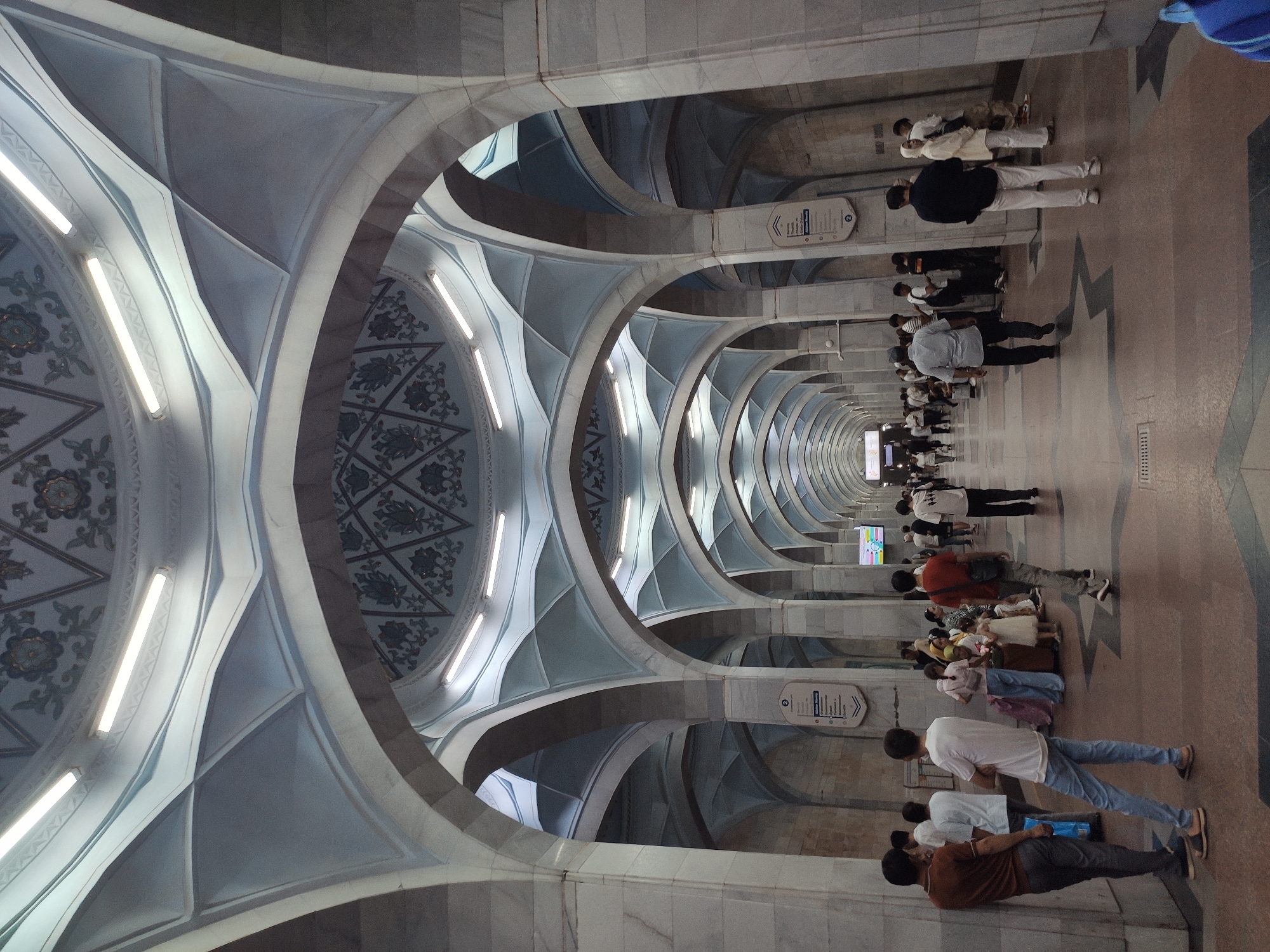 This is a photograph of the interior of the Mirzo Ulugbek metro station in Tashkent, Uzbekistan. The station features a long, arched hallway lined with marble columns. The ceiling is a prominent architectural element, with intricate geometric patterns and decorative motifs in shades of blue and white. Fluorescent lights are embedded in the ceiling, illuminating the space.
A number of people are present in the hallway, standing, walking, and congregating in small groups. They are diverse in age and attire. Some individuals are waiting, while others appear to be passing through. The floor is a patterned tile, with a dark reddish-brown hue and lighter geometric inlays. Signage is visible on some of the columns, indicating information or directions. The overall impression is one of a clean, well-lit, and architecturally distinct public space.