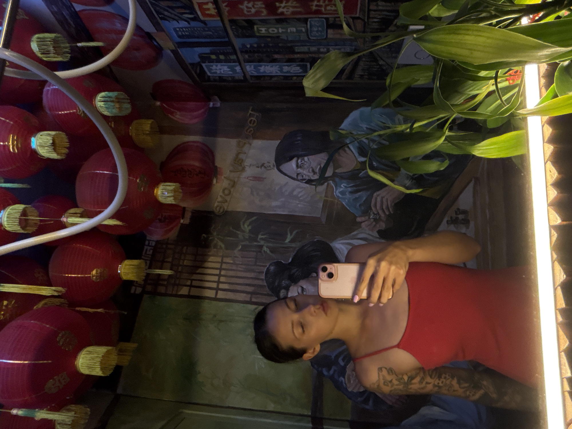 The image shows a mirror selfie taken by a woman wearing a red dress. The mirror reflects a mural depicting two Asian figures. The background behind the mural features red lanterns and signs with Asian characters. Green leafy plants are visible in the upper right corner of the image. The mirror is framed by a bright, narrow light source. The image was taken in North Kuta, Indonesia.