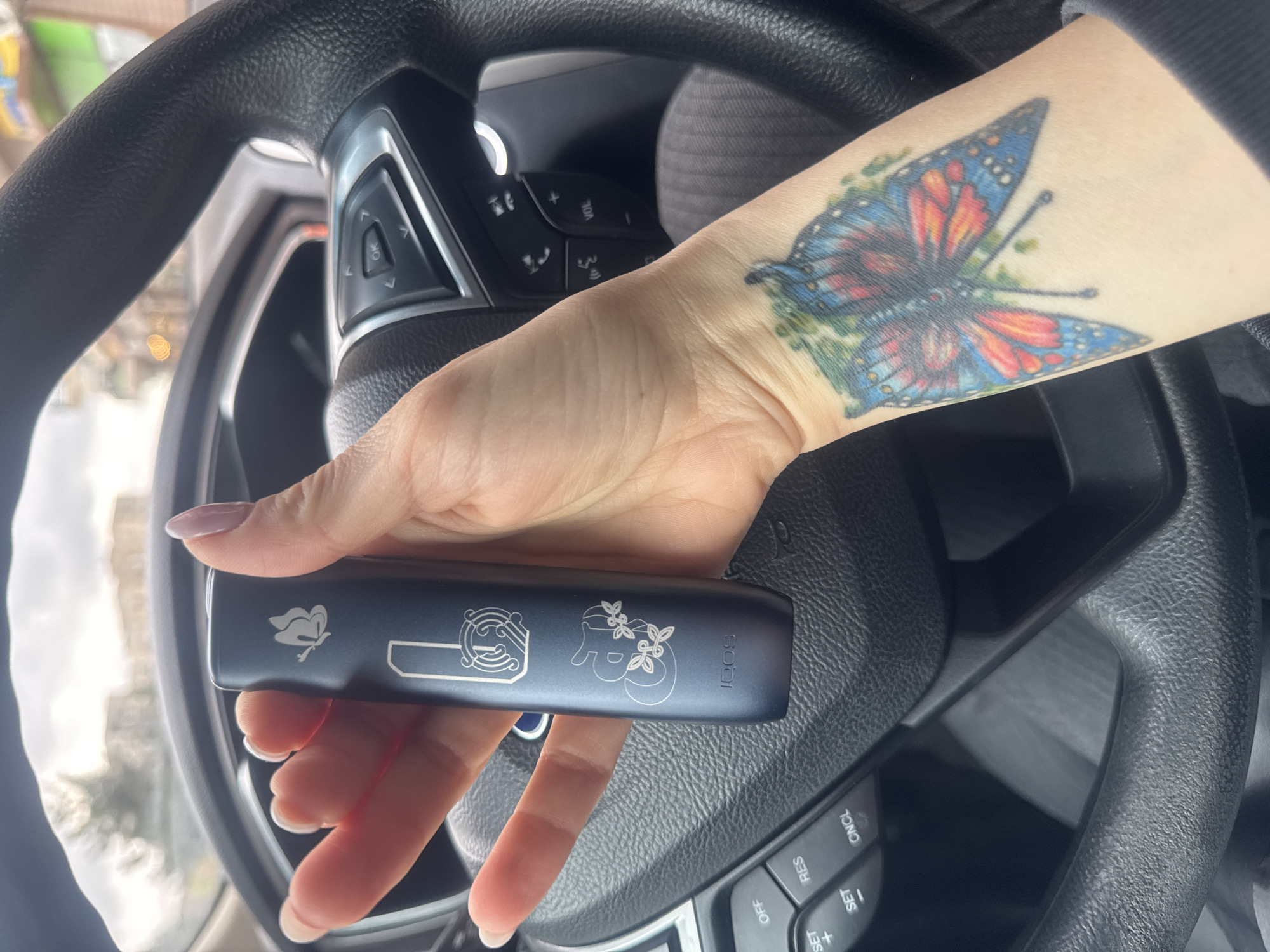 A close-up, eye-level shot captures a person's arm and hand inside a car, holding a dark blue electronic device. The hand has light-colored, manicured nails. Prominently displayed on the forearm is a vibrant butterfly tattoo, intricately designed with shades of blue, orange, red, and green.
The electronic device, possibly a vape or heated tobacco product, features several etched designs, including a stylized butterfly, abstract patterns, and a cluster of berries or leaves. The brand name "IQOS" is clearly visible on the side of the device.
The background shows parts of a car's black steering wheel, indicating the setting is inside a vehicle. Control buttons are visible on the steering wheel, displaying text such as "OK", "VOL", "ONCL", "RES", "SET", "+", and "-". The exterior through the car window is blurred but suggests daylight conditions, with light filtering in from an outdoor environment that hints at buildings or natural elements. The activity is simply a person holding the device while in a car. The image is described as being from Tbilisi, Georgia.