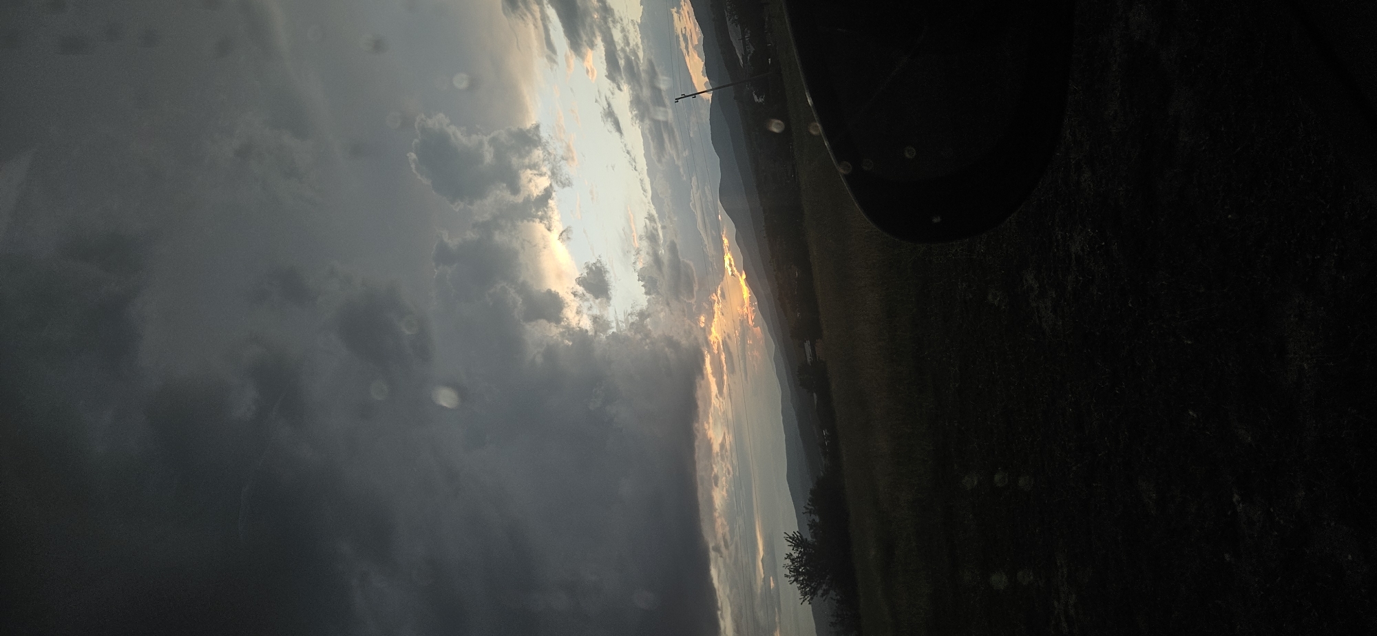 The image captures a dramatic outdoor landscape, likely at sunrise or sunset, showcasing a stark contrast between heavy storm clouds and a brightly illuminated horizon. The sky on the left is dominated by dense, dark grey clouds, while towards the right, the clouds lighten, revealing a brilliant golden and orange glow where the sun breaks through. This light illuminates patches of the sky and the distant horizon.
Across the mid-ground, a range of mountains stretches, appearing as dark silhouettes against the vibrant sky. Below the mountain range, a dark, open field occupies the foreground, with some sparse foliage or small trees visible. A single utility pole stands vertically near the center-right of the horizon line. In the lower right corner of the frame (appearing in the upper right due to the image's rotation), a dark, circular object, possibly part of a vehicle's tire or fender, intrudes into the view. Scattered across the entire image, particularly noticeable against the darker areas and sky, are numerous small, translucent circular artifacts, which appear to be water droplets or specks of dust on a lens or window.
The scene conveys the raw power of nature during a weather transition, with the interplay of light and shadow creating a powerful atmosphere. The location context is Momin Prohod, Bulgaria. No people or discernible text are visible in the image.