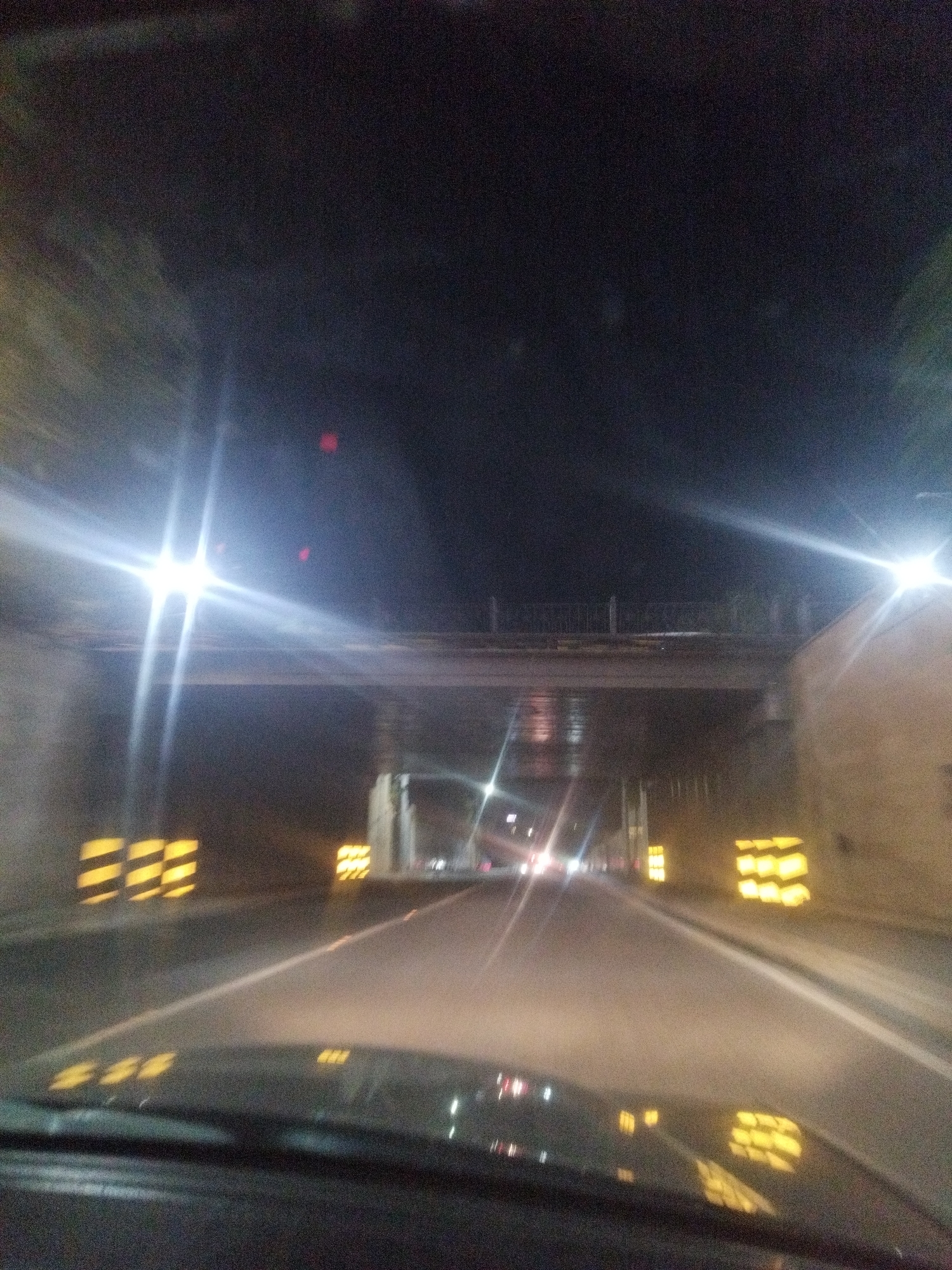 The image depicts a night-time view from the perspective of someone driving a vehicle on a road that appears to be under a bridge or overpass. The road is paved and has lane markings. On either side of the road, there are reflective yellow and black barriers or signs. The environment is dark, with artificial lighting from streetlights and vehicle headlights creating glare and reflections. The structure above is dimly lit, and there is a hint of foliage on the sides of the road. The overall impression is that of an urban or semi-urban setting at night, possibly a highway or major road. The image does not contain any discernible people or specific landmarks that would allow for precise location identification within Shymkent, Kazakhstan.