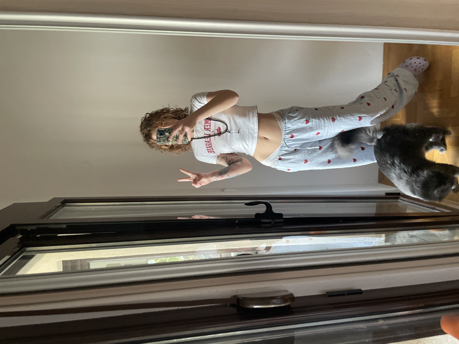 The image portrays an interior scene within a residence in Boreti, Montenegro. A young adult female, reflected in a mirror, is depicted taking a selfie with a smartphone held to her face, obscuring her features. She has light brown curly hair, wears a white cropped t-shirt with partial red text, and light blue and white striped pajama pants with red heart patterns and white lace trim at the waist. A dark bead necklace is visible around her neck. Tattoos are present on both forearms. Her left hand is raised, forming a peace sign. A single light-colored, patterned slipper is worn on her right foot (left in the reflection). On the wooden floor, a grey domestic cat with a fluffy tail stands near her legs. To the left, a multi-pane glass door or window with a dark frame and handle reveals a portion of an exterior wall and sparse greenery. The room features plain white walls and warm-toned wooden flooring.