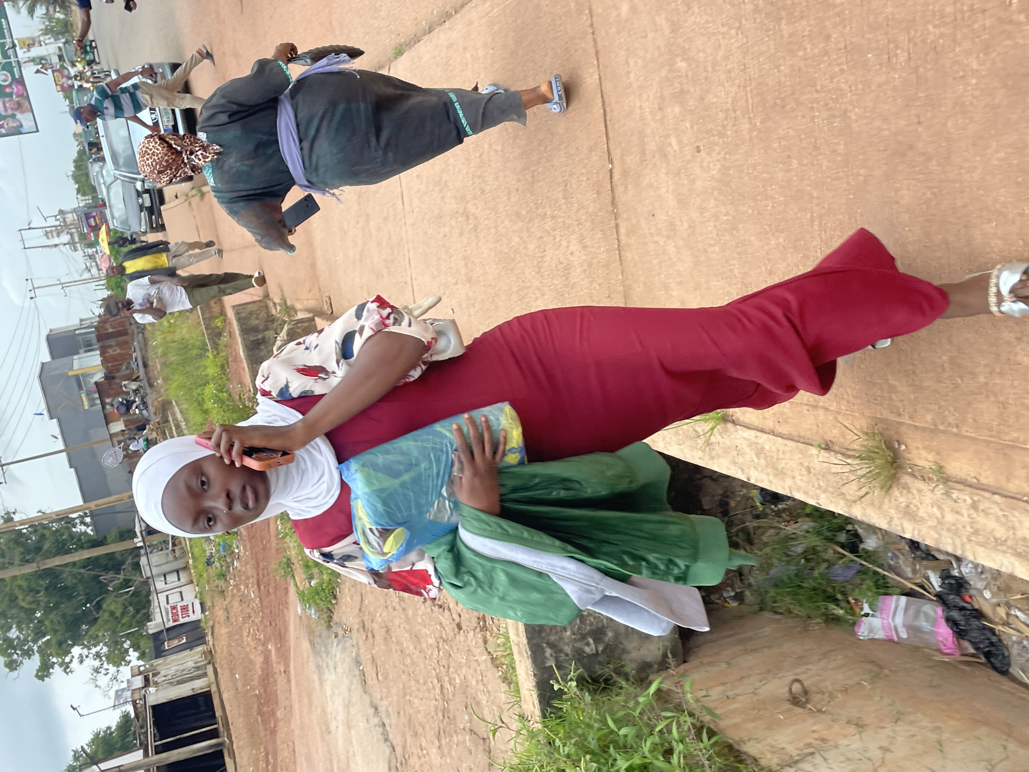 A young Black woman wearing a white hijab and a maroon dress is walking on a paved path in Jos, Nigeria. She is holding a blue plastic bag and a green jacket in her left arm, while her right hand is raised to her ear, holding a pink smartphone. She is looking towards the camera with a neutral expression. In the background, other people and vehicles are visible on a street. A gutter filled with trash is located to the left of the paved path. The area appears to be an urban setting with buildings and a road. The sky is overcast.