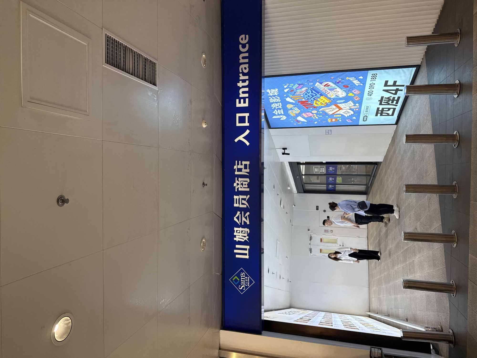 The image shows the entrance to a Sam's Club store, likely in Tianhe District, China, as indicated by the Chinese text on the signage. Above the entrance, a prominent blue sign reads "Sam's Club" in English and Chinese characters, followed by "入口 Entrance". To the right of the main entrance, a large digital screen displays colorful advertisements for a cinema, featuring the text "金逸影城" (Jinyi Cinema), a phone number "400-090-1888", and "西座4F" (West Block 4F).
In front of the entrance, a walkway is lined with several polished metal bollards. Inside the entrance area, three people are visible. On the left, a woman in a white shirt and black pants stands looking towards a large illuminated display on the left wall. In the center, a man and a woman, both dressed casually, are standing and conversing. The man is wearing a light-colored t-shirt and dark pants, while the woman is in a light blue shirt and dark pants. The interior walls of the entrance area are white. The ceiling is a tiled grid with recessed lighting and a ventilation grate.