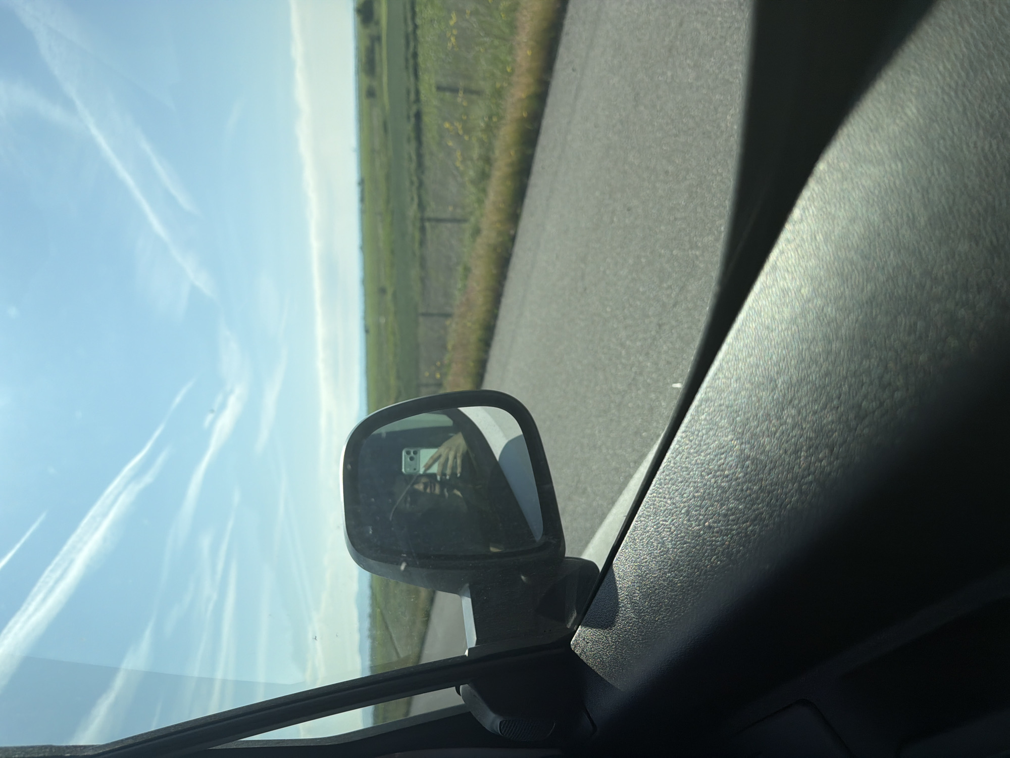 The image shows a view from inside a vehicle, with a side mirror visible. The reflection in the side mirror captures a person holding a smartphone, presumably taking the photo. Outside the vehicle, there is a road with fields and a blue sky in the background. No explicit information suggests the location is in Загорци, Bulgaria.