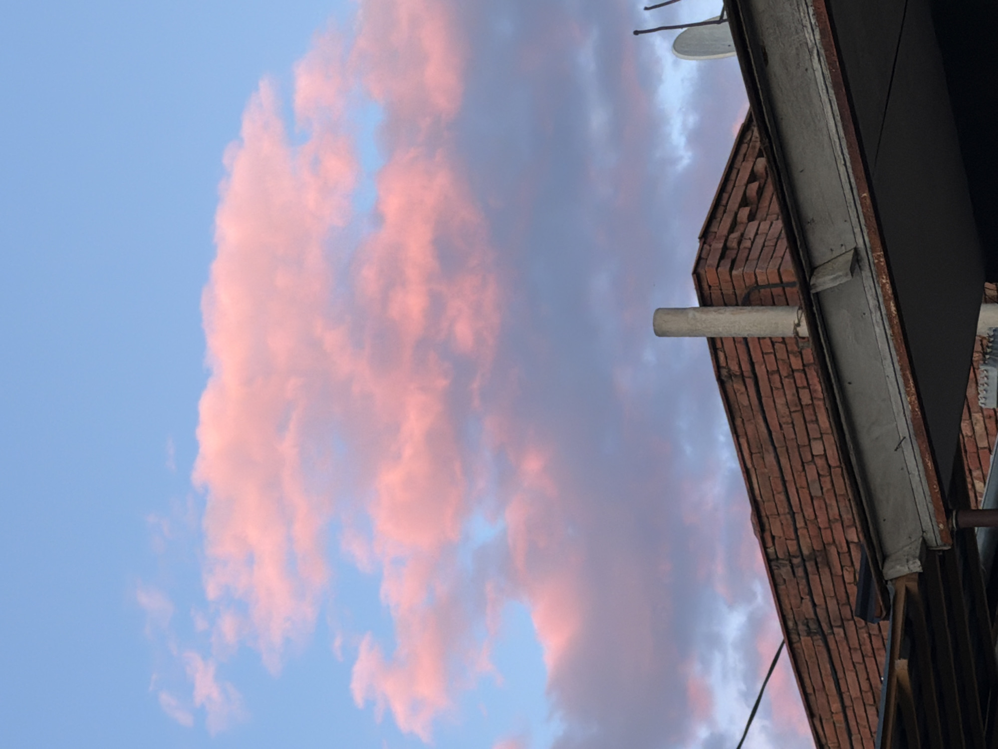 The image shows a view of the sky and a building in Tbilisi, Georgia. The sky is a light blue color with pink and gray clouds. The building is made of red brick and has a gray pipe extending from its side. A satellite dish is also visible on the roof. The angle of the shot suggests it was taken from a low vantage point, possibly from street level.
