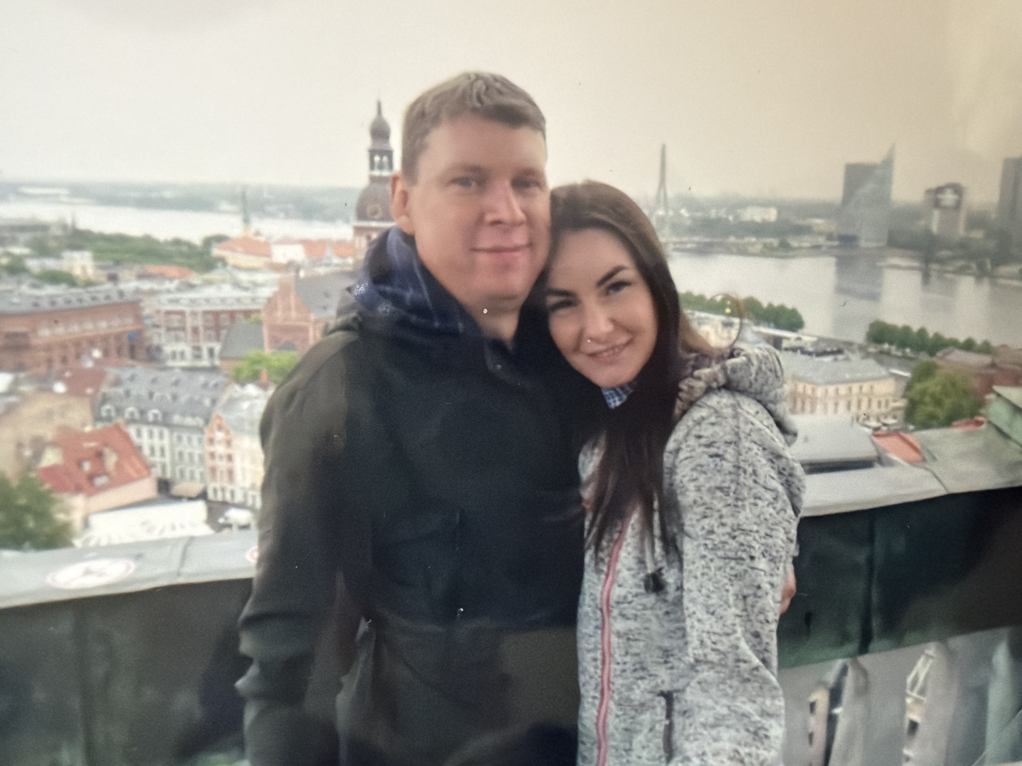 A man and woman are posing closely together for a photograph with a cityscape in the background. The man is wearing a dark jacket with a hood, while the woman is wearing a grey hooded jacket. The cityscape behind them features a river, buildings with distinct architectural styles, and a tower with a spire. The sky appears overcast. The location is Riga, Latvia.