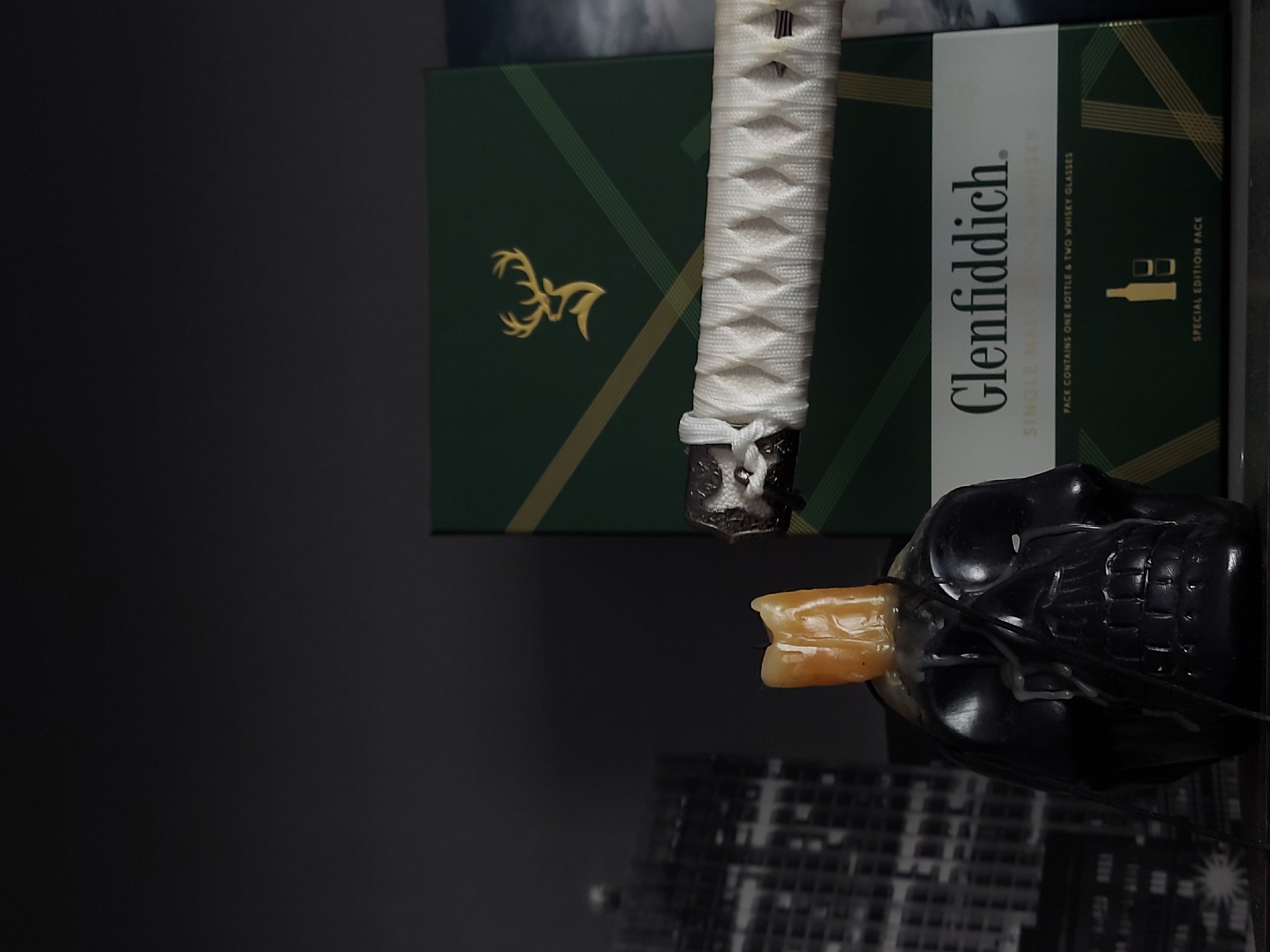 The image shows a still life composition featuring a bottle of Glenfiddich whisky, a katana sword handle, and a skull. The Glenfiddich box is green with gold accents and the brand name prominently displayed. The katana handle is white with a dark-colored pommel. A black skull with an orange object protruding from its mouth sits in front of the Glenfiddich box. The background has a blurry cityscape pattern. There is no information to identify if this scene is in Izhevsk, Russia.