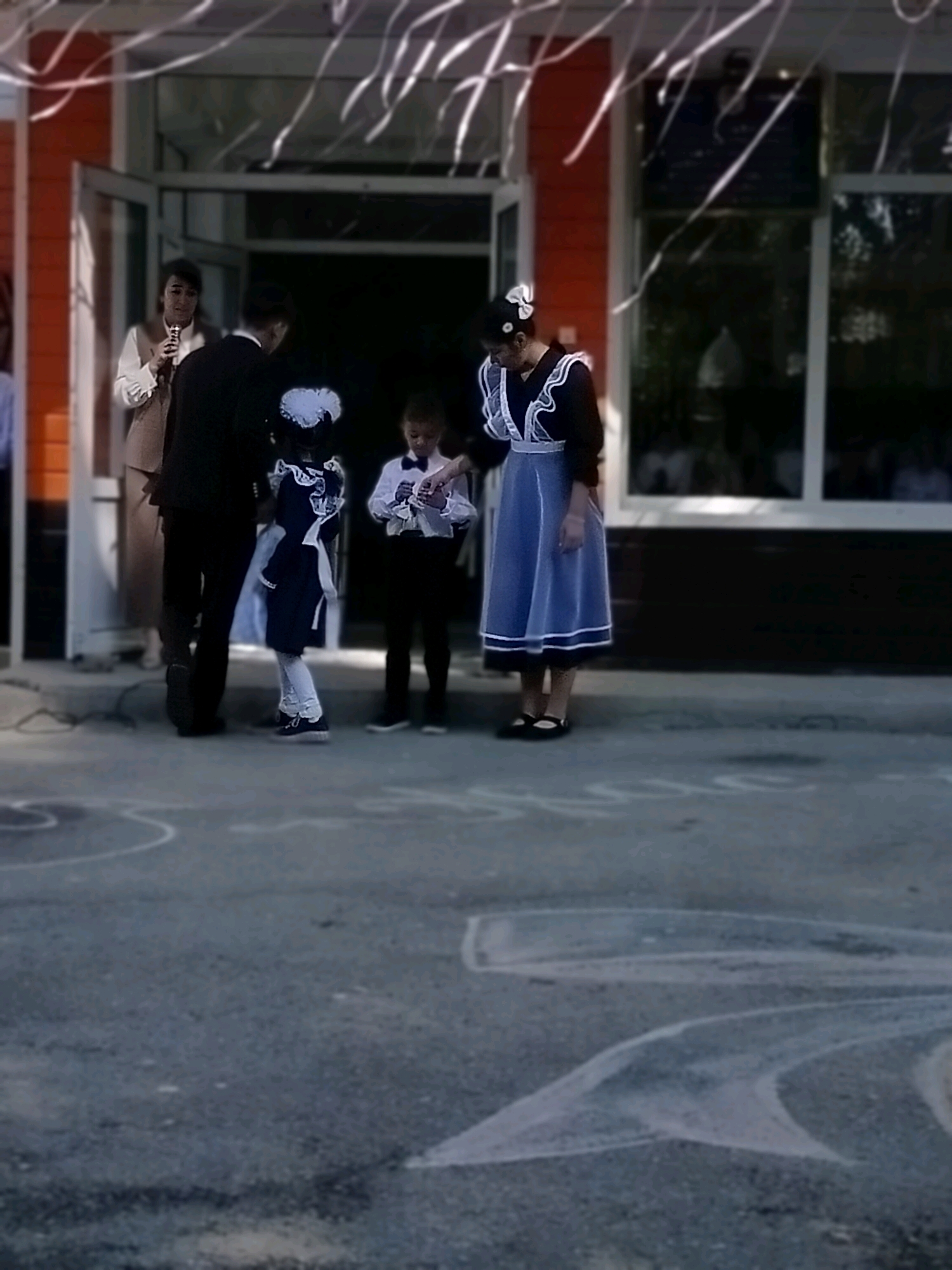 The image captures an outdoor gathering in front of a building with orange siding and large glass doors, possibly in the city of Kölkent, Kazakhstan. Several individuals are present. In the background, a woman in a white blouse and light brown vest stands holding a microphone. To her right, a man in a dark suit faces away from the camera. In the foreground, two children in school uniforms are visible. One child, in a dark dress with a white pinafore and bow, stands facing the viewer. Another child, in a white shirt with a bow tie and dark pants, is also present. The ground is paved and features white chalk markings. Decorative white ribbons are suspended in the air above the scene.