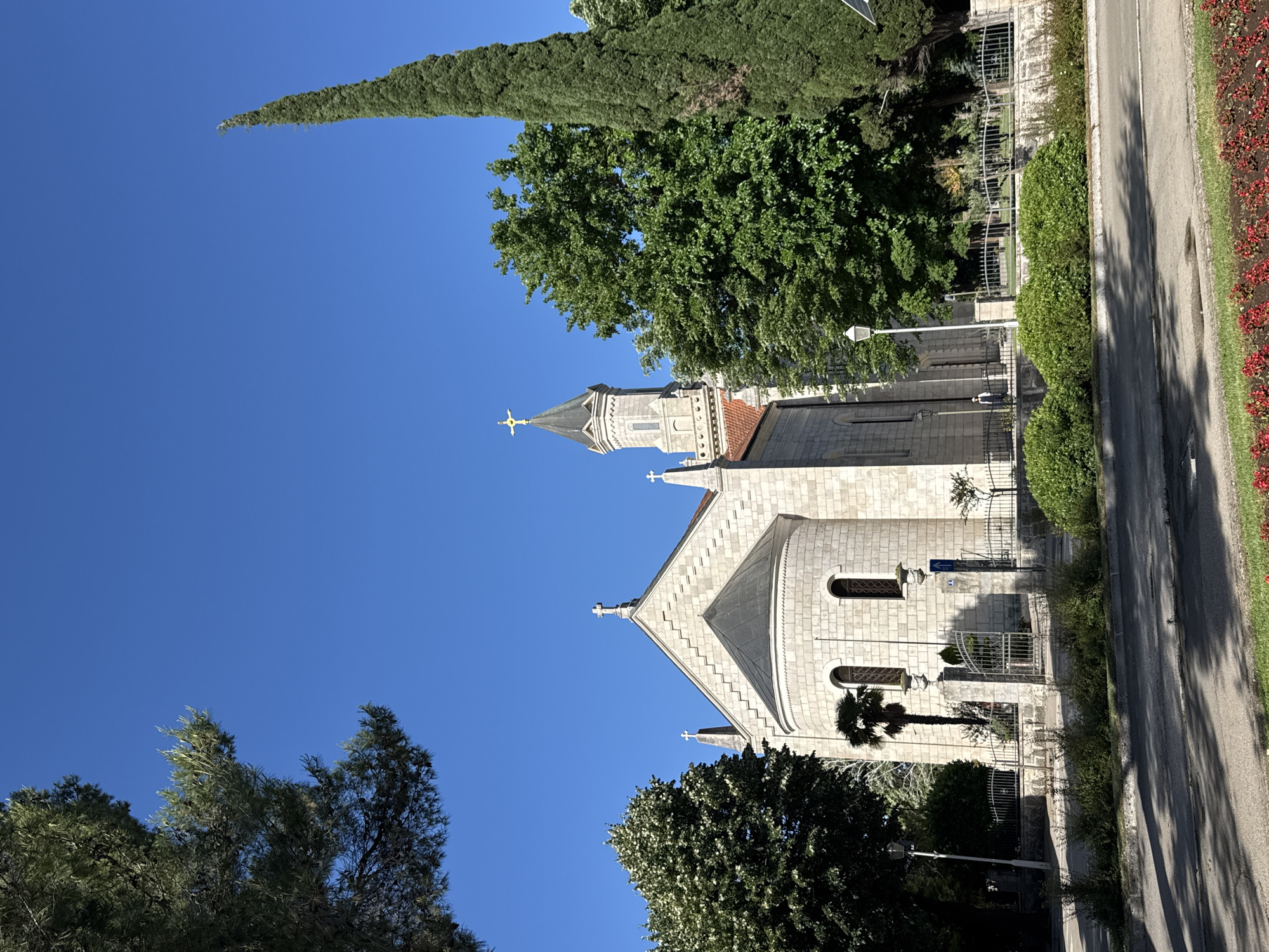 The image depicts the Church of the Transfiguration of Our Lord in Trebinje, Bosnia and Herzegovina. The church, constructed from light-colored stone, features a prominent bell tower with a golden cross at its apex. A semicircular apse is visible on the left side of the building, adorned with arched windows. Two smaller crosses are also present on the church's roof.
The church is situated on a slightly elevated plot, bordered by a metal fence and manicured hedges. A paved road runs in front of the church, with a sidewalk and a strip of red flowers. Several mature trees, including a tall cypress tree on the right and a broad leafy tree on the left, frame the scene. The sky is clear and a vibrant blue. A single person, dressed in dark clothing, is walking on the sidewalk behind the fence.