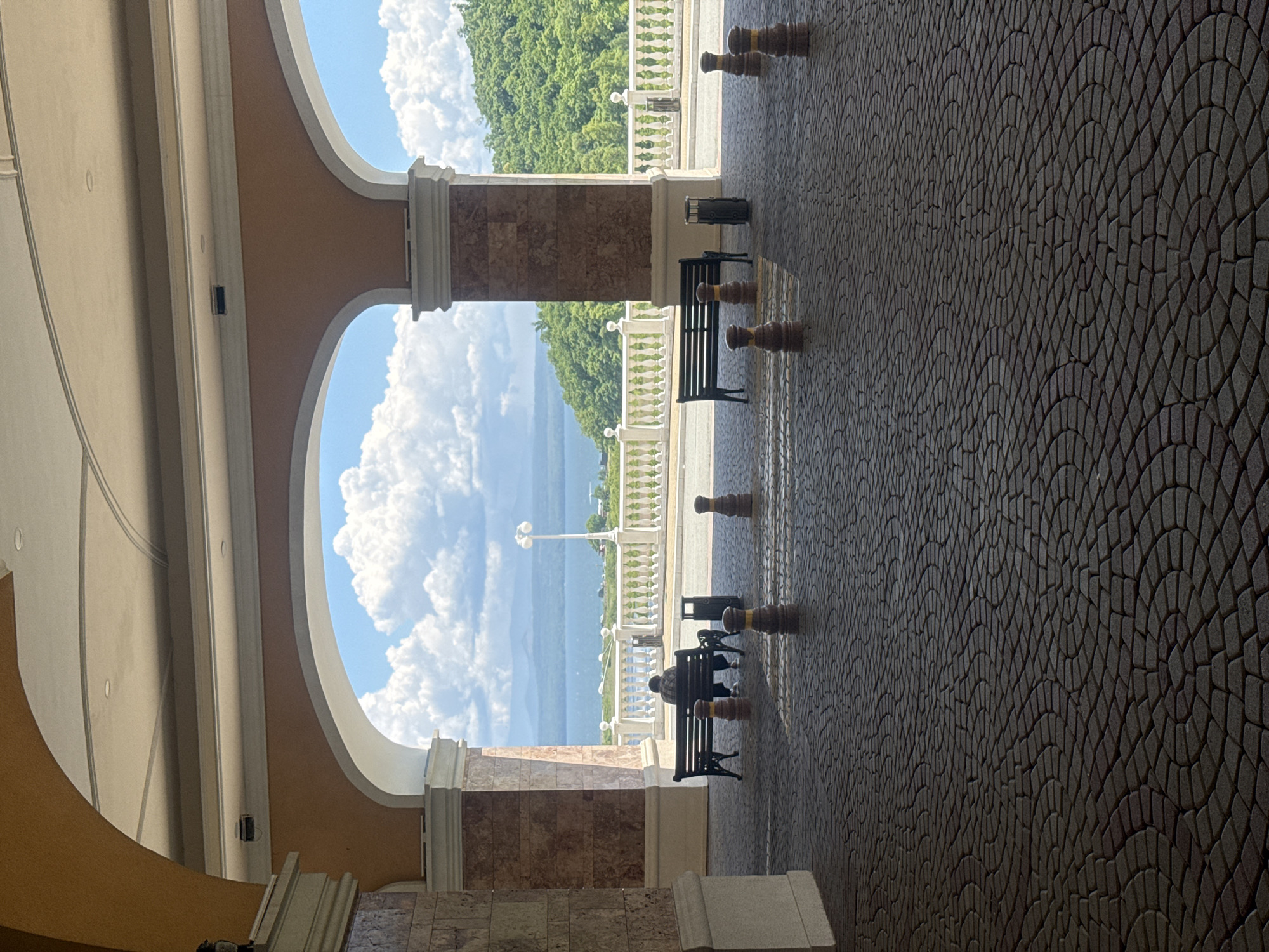 The image depicts a view through an arched walkway towards a body of water and a lush green hillside. In the foreground, a person is seated on a bench, facing away from the camera, looking out at the scenery. The ground is paved with interlocking stones arranged in a circular pattern. Several bollards are also visible on the paving. Beyond the walkway, there is a white railing overlooking the water, with trees and hills in the distance. The sky is filled with dramatic white clouds. The scene appears to be outdoors in a well-maintained area, possibly a public space or resort. There is no indication of the specific location within Shkotovo, Russia.