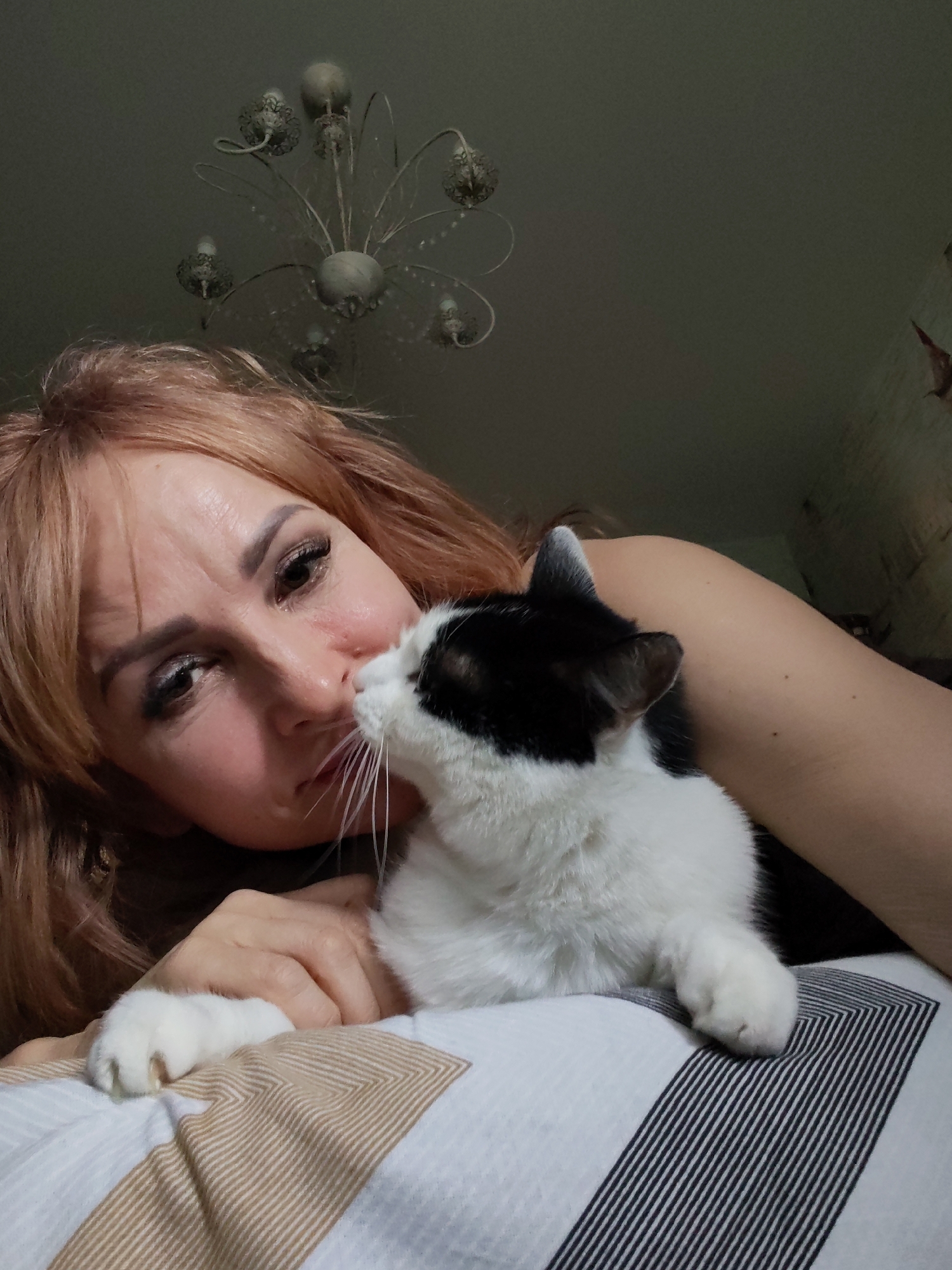 This close-up indoor image, taken in Chaikovsky, Russia, features a woman and a black and white cat engaged in a tender interaction. The woman, with reddish-blonde hair, is lying down, her head resting on a striped pillow. She wears eye makeup, including dark eyeliner, eyeshadow, and filled-in eyebrows, and her expression is soft and appears affectionate as she looks towards or slightly past the cat.
The cat, predominantly white with black patches on its head, ears, and back, is positioned very close to the woman's face. Its nose is touching or nearly touching the woman's nose, suggesting an intimate moment of sniffing or a "cat kiss." One of the cat's white paws, with visible claws, rests gently on the striped pillow, while another paw is also visible.
In the background, a decorative, multi-armed chandelier hangs from a light-colored ceiling, indicating an indoor setting, likely a home, possibly a bedroom. The lighting is subdued, suggesting ambient light rather than bright daylight, hinting at an evening or nighttime scene. The overall atmosphere conveys warmth, comfort, and a strong bond between the woman and her feline companion. There is no visible text in the image.