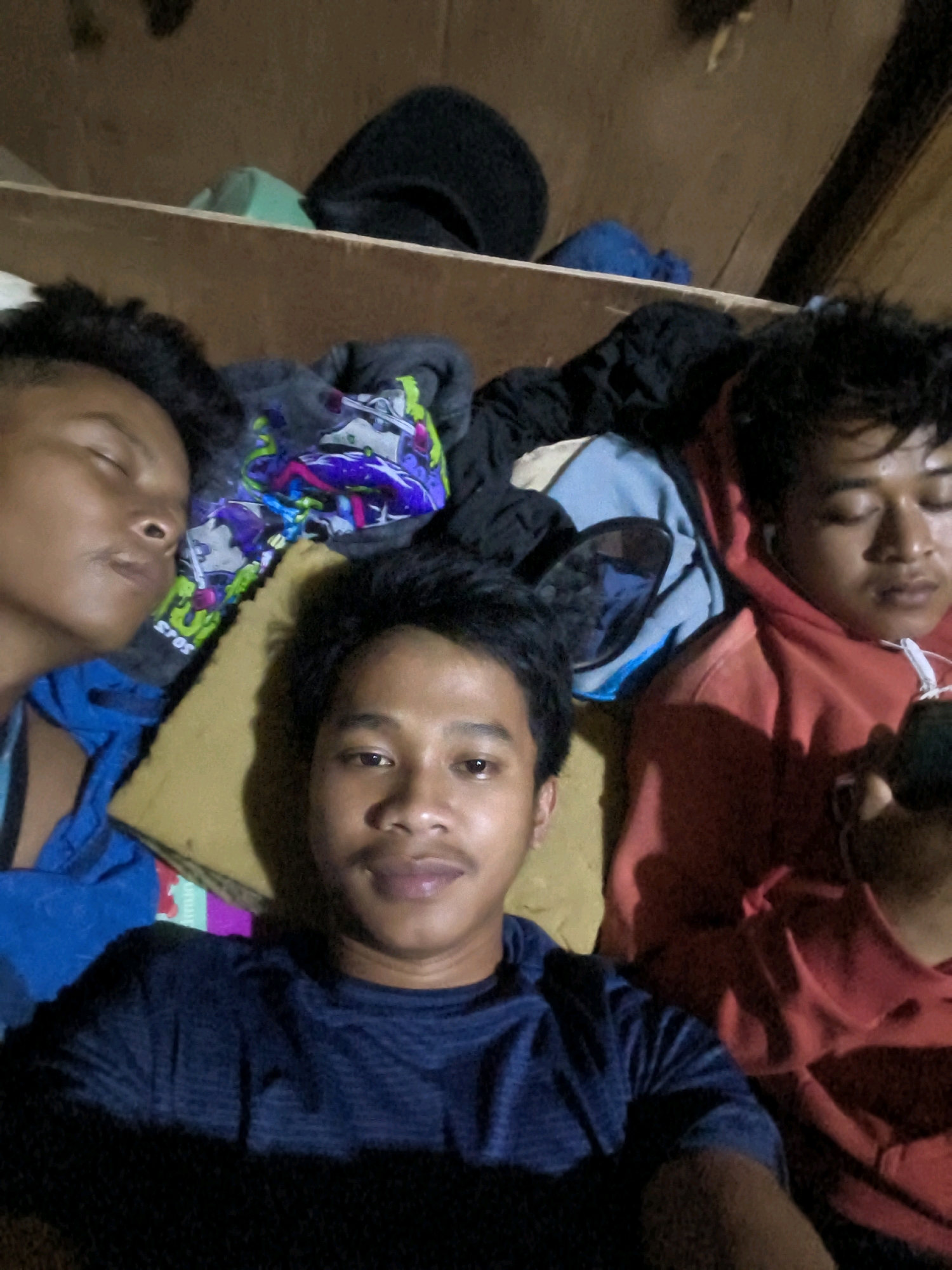 The image is a selfie of three young men in what appears to be a dimly lit room, possibly a dormitory or a shared living space, in Bekasi, Indonesia. Two of the men are lying down and appear to be asleep, with their eyes closed. One of these sleeping individuals is wearing a blue shirt, and the other is wearing a red hoodie with white earbuds. The third man is in the foreground, looking directly at the camera with his eyes open and a slight smile. He is wearing a dark blue t-shirt. The background shows some clothing items and a wooden shelf. The overall lighting is low, suggesting it might be nighttime or the room is poorly lit. The context suggests a casual and personal moment captured by the individuals themselves.